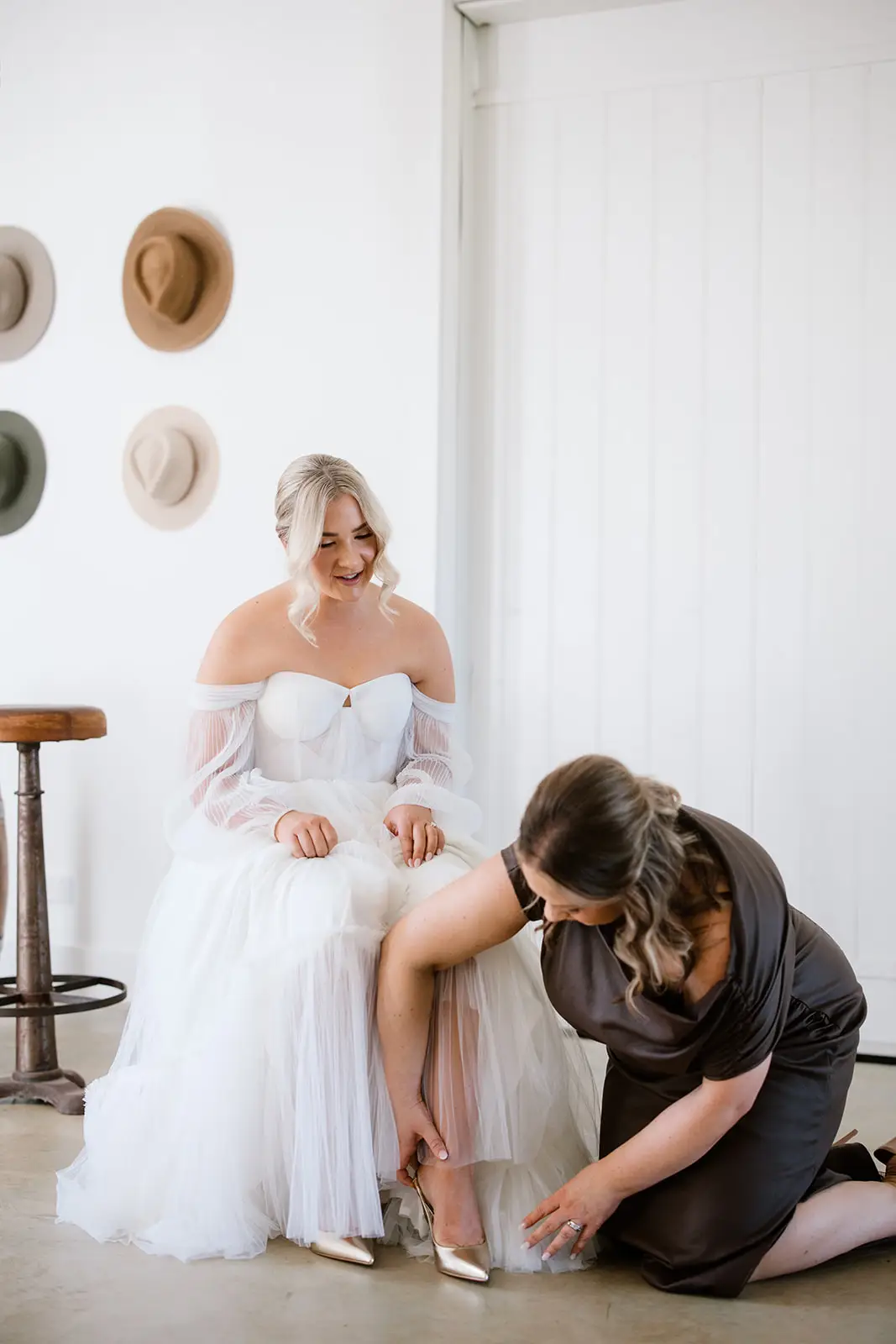A bride in an off-the-shoulder pleated tulle wedding dress sits as her bridesmaid in a dark satin dress helps her put on metallic gold pointed-toe pumps.