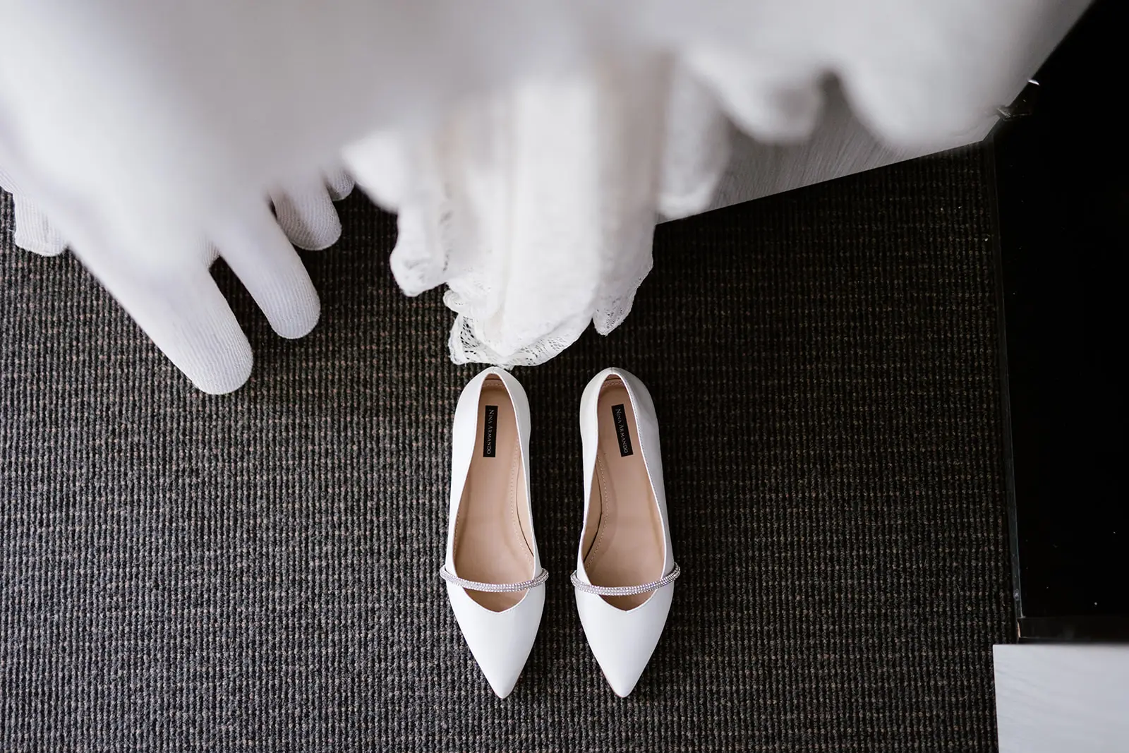 Overhead detail shot of Nina Armando white pointed-toe bridal flats with a thin crystal-embellished Mary Jane strap. The shoes are placed neatly on a dark grey textured carpet next to the lace hem of a white wedding dress.