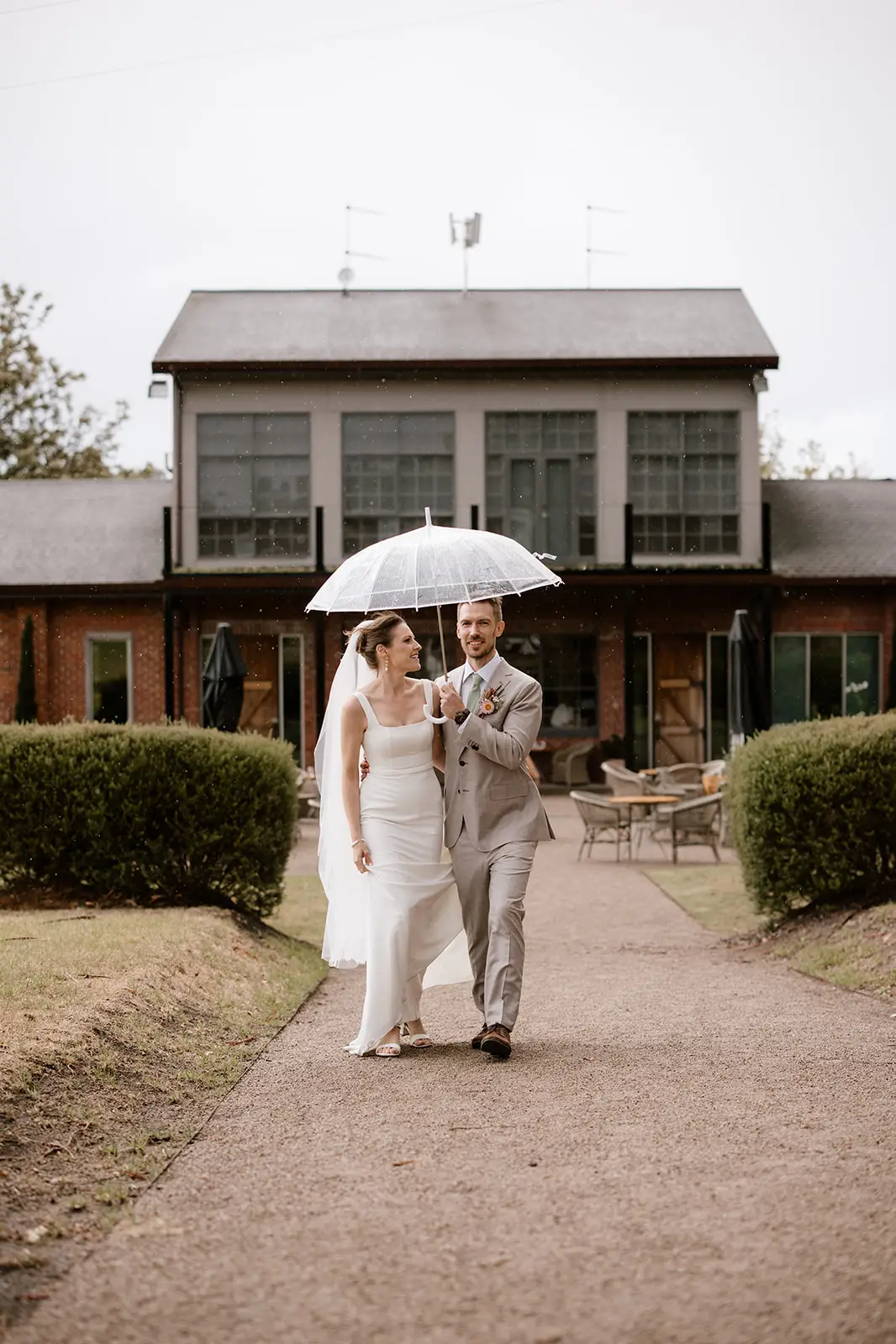 A bride in a sleek white square-neck wedding gown and a groom in a light grey suit walking together under a clear umbrella during a light rain. They are walking on a textured asphalt path at an estate-style venue, with the bride lifting her skirt to reveal white strappy sandals.