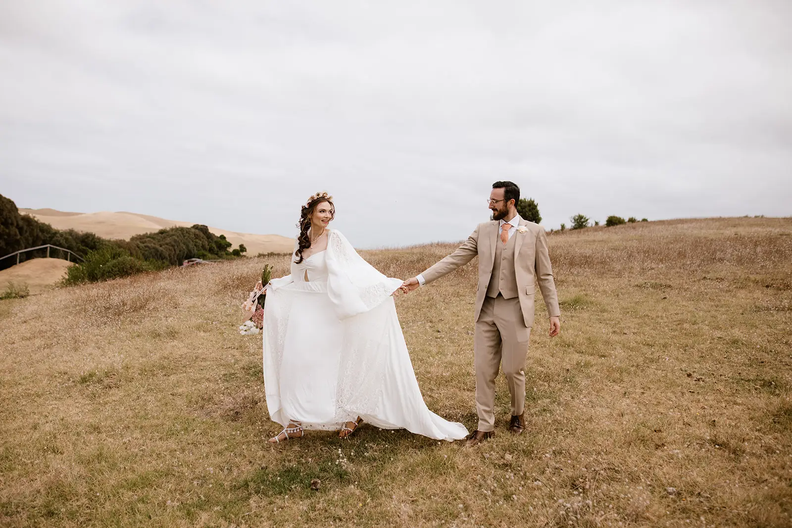 Bride wearing white boho tassel sandals walks through a dry grassy field, holding her husband's hand. The flat bridal sandals feature ankle wraps and decorative tassels, making them ideal footwear for an outdoor meadow wedding.