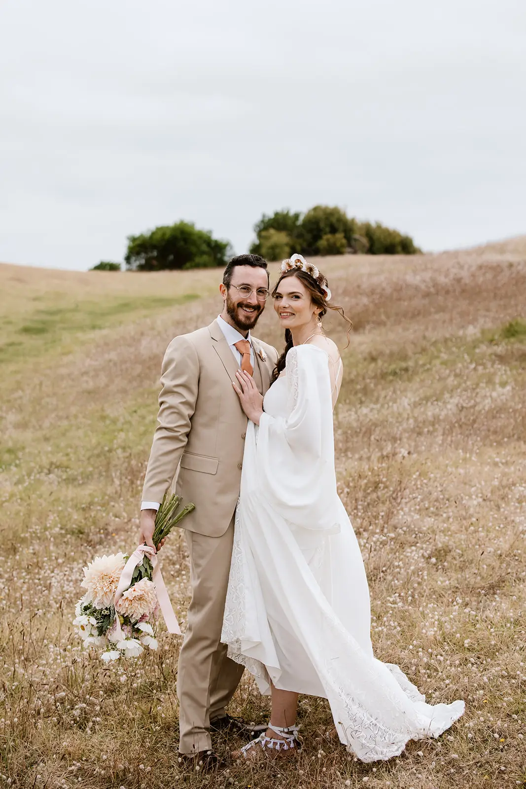 Close-up profile shot of a bride in a white lace-trimmed wedding dress and boho tassel sandals standing in a grassy meadow with the groom. The bride's flat white sandals feature decorative tassels and ankle straps, providing a practical yet stylish footwear option for a rural hillside wedding.