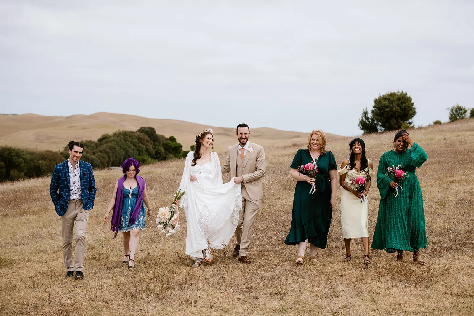 A bride, groom, and their bridal party walk across a dry, grassy meadow. The bride lifts her white dress to reveal flat boho tassel sandals, while the bridesmaids wear a mix of green and yellow dresses with sensible block heels and sandals suited for the outdoor terrain.