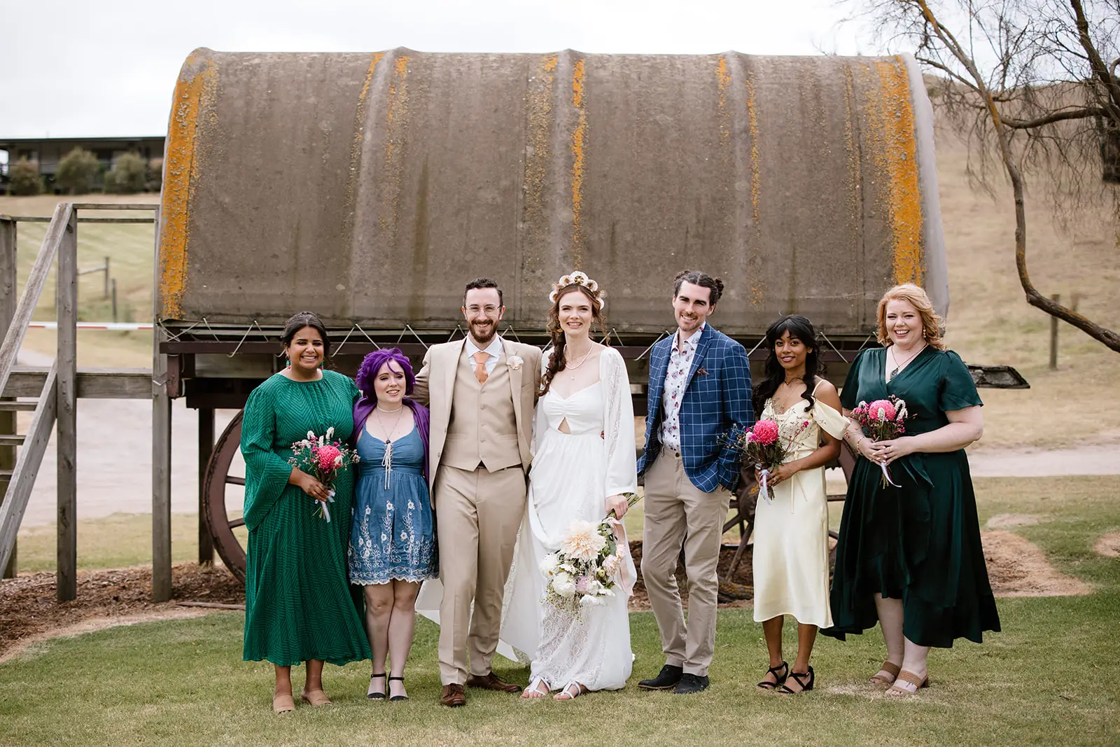 A full-length group shot of a wedding party standing on grass in front of a rustic wagon. The bride is wearing flat white sandals with tassels. The bridesmaids wear a variety of outdoor-appropriate footwear including block-heel sandals and flats in tan and black.