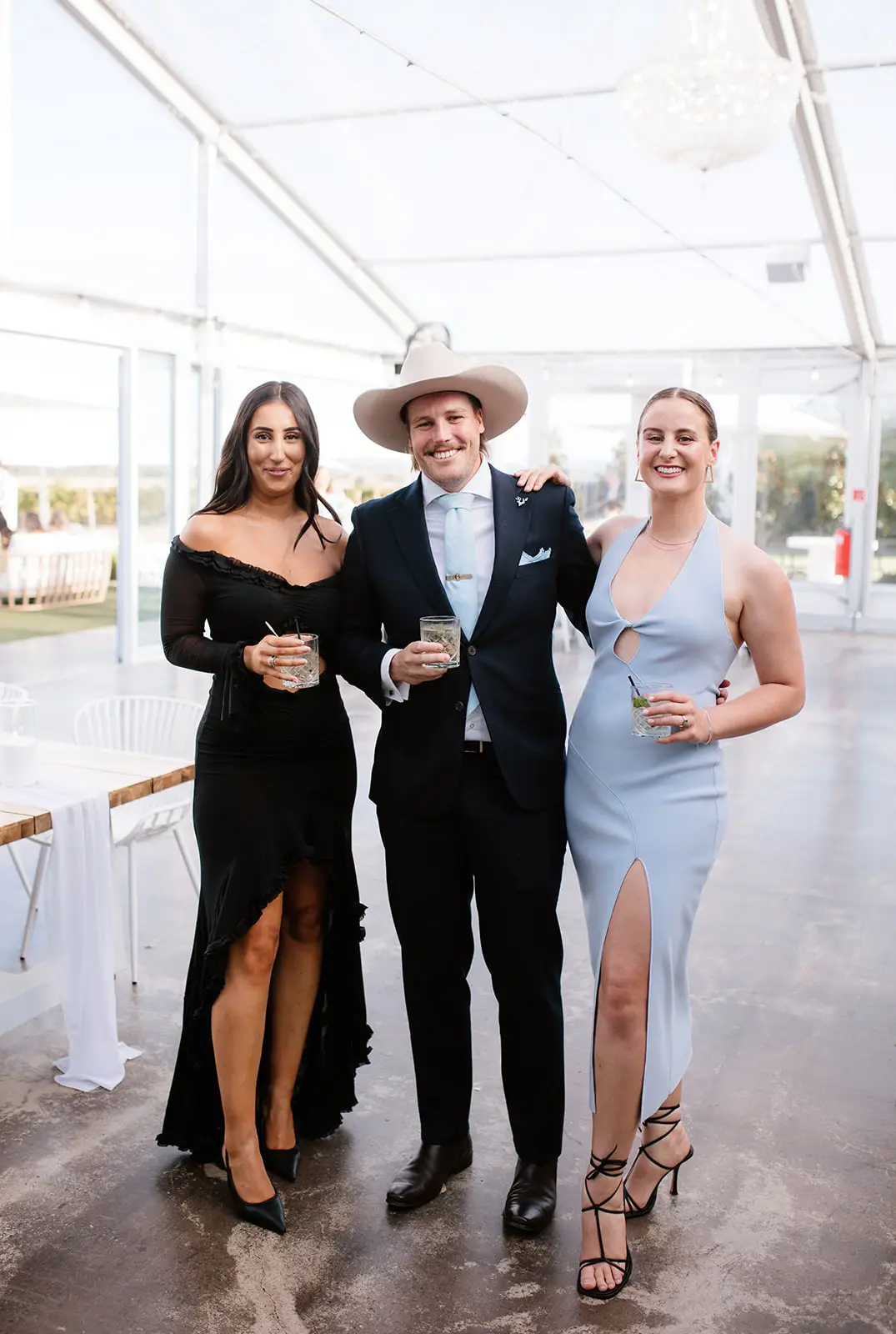 A wedding guest in a light blue halter midi dress wearing black lace-up dressy sandals with a high stiletto heel, standing on a concrete floor in a marquee.