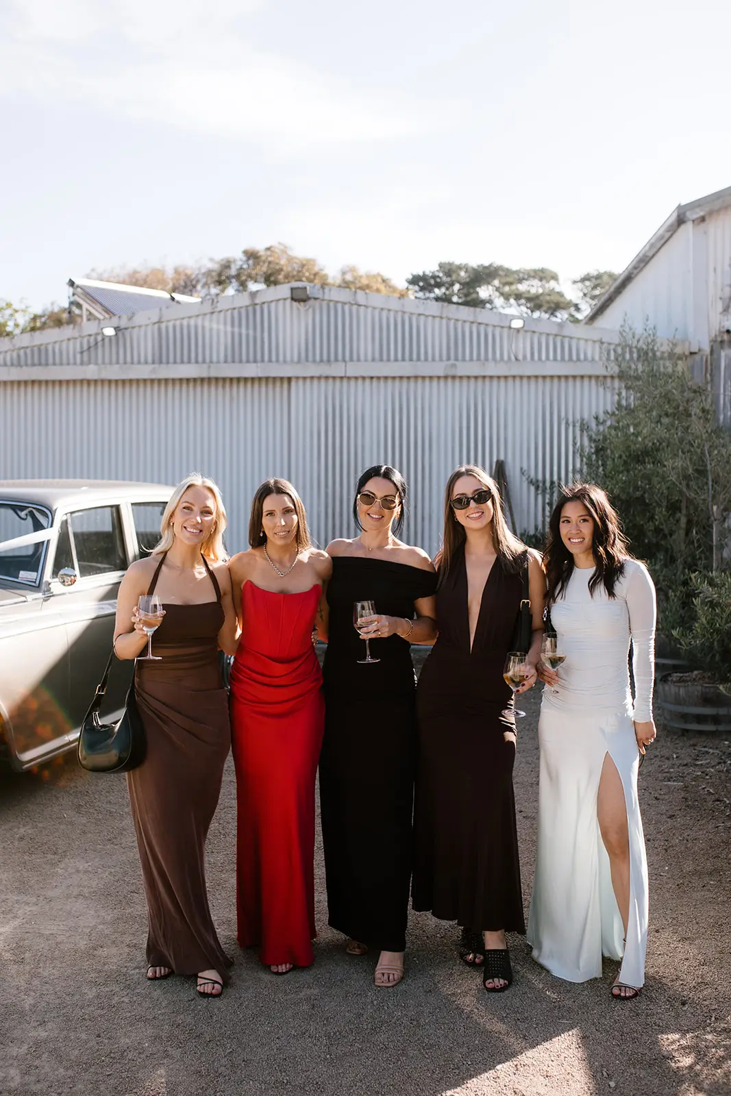 A group of five wedding guests in various formal gowns—brown, red, black, and white—standing on a gravel driveway, showcasing different styles of dressy sandals including strappy flats and block heels.