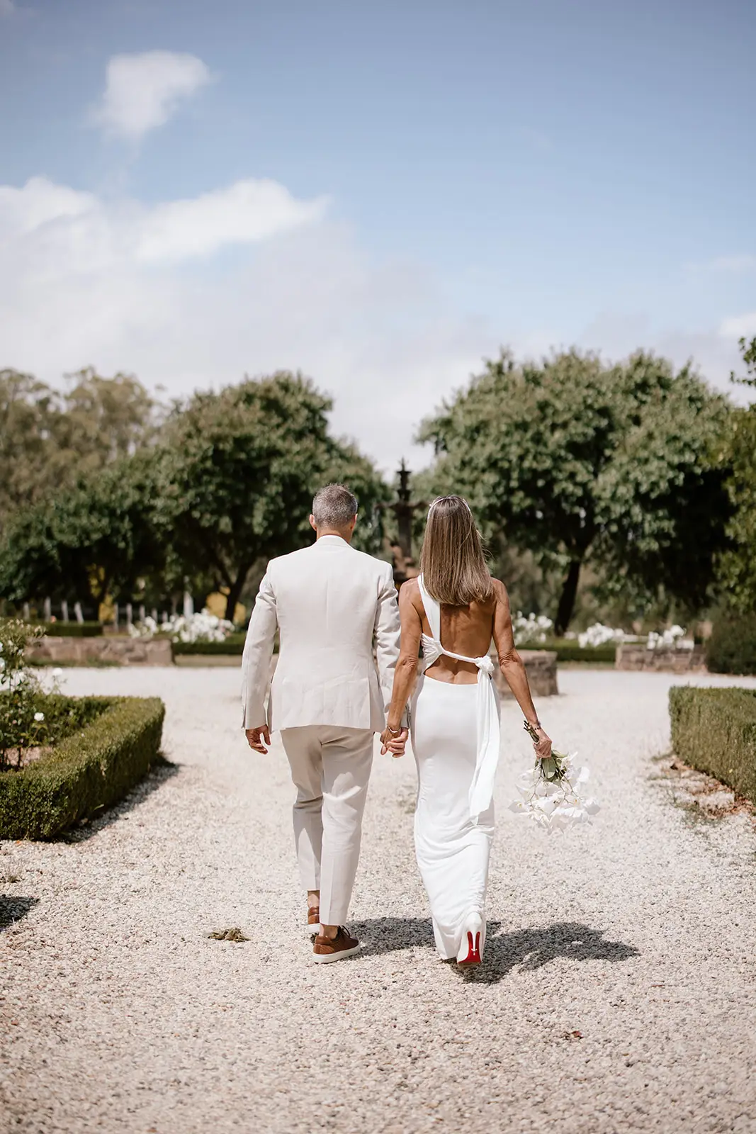 A bride and groom walking away from the camera on a light-colored gravel path. The bride wears a sleek, backless white wedding dress and iconic Christian Louboutin high heels with signature red soles. The groom wears a neutral-toned linen suit and brown leather sneakers. They are walking toward a lush green garden under a bright blue sky on the Mornington Peninsula.
