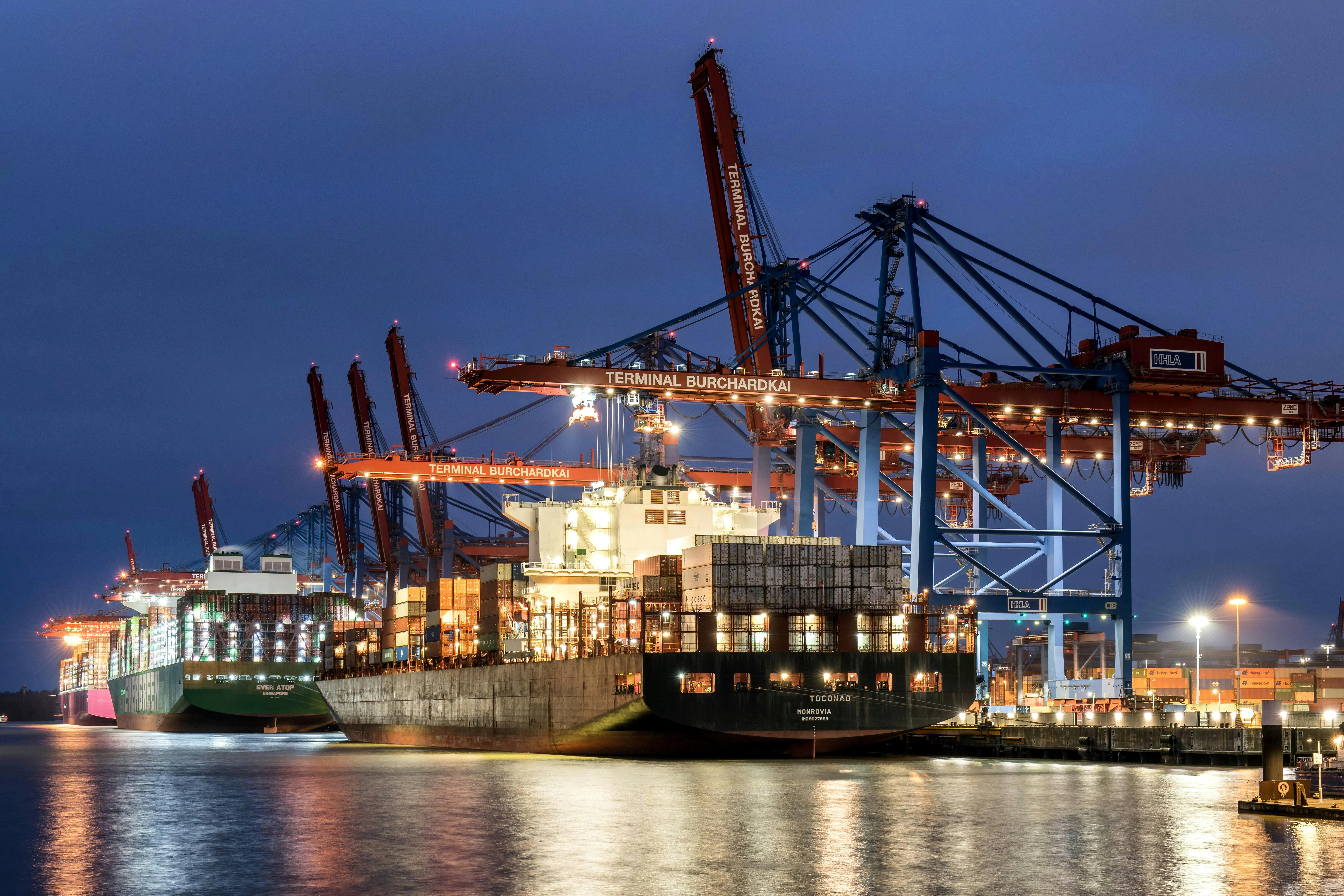 A shipyard with large cranes and shipping containers photographed in the evening