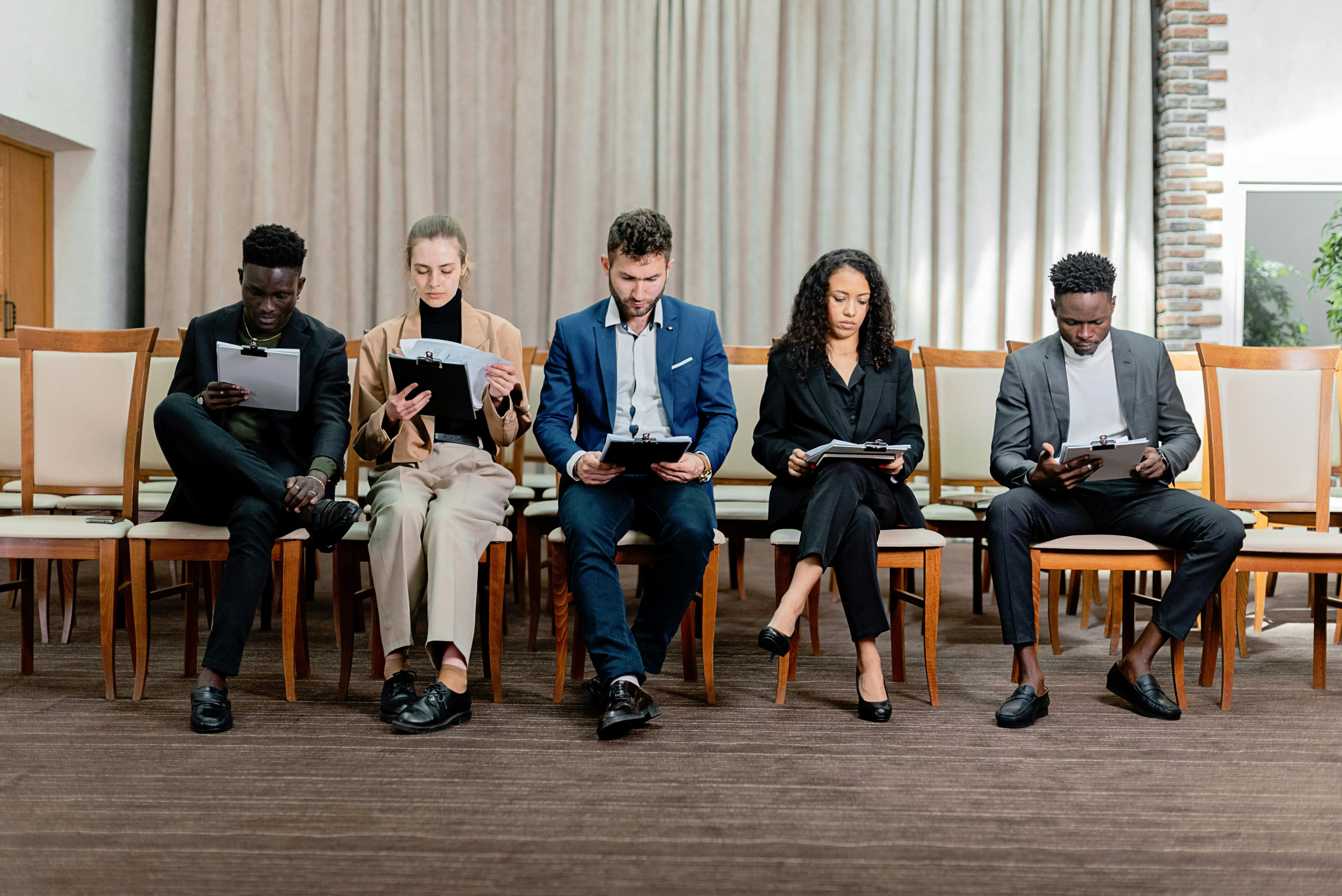 Five people sitting next to each other on chairs holding either a book or some papers getting ready for an interview