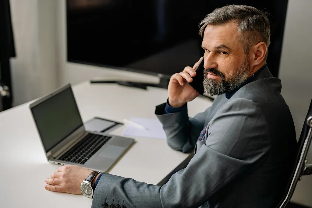 A man wearing a suit talking on the phone with a laptop in front of him while sitting at a table