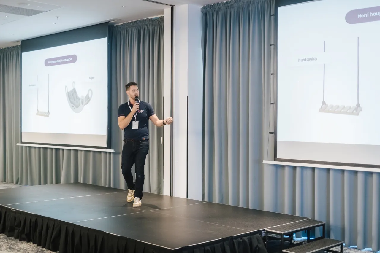 Muž držící mikrofon a prezentující na pódiu s projekcí obrázků houpaček na pozadí.
