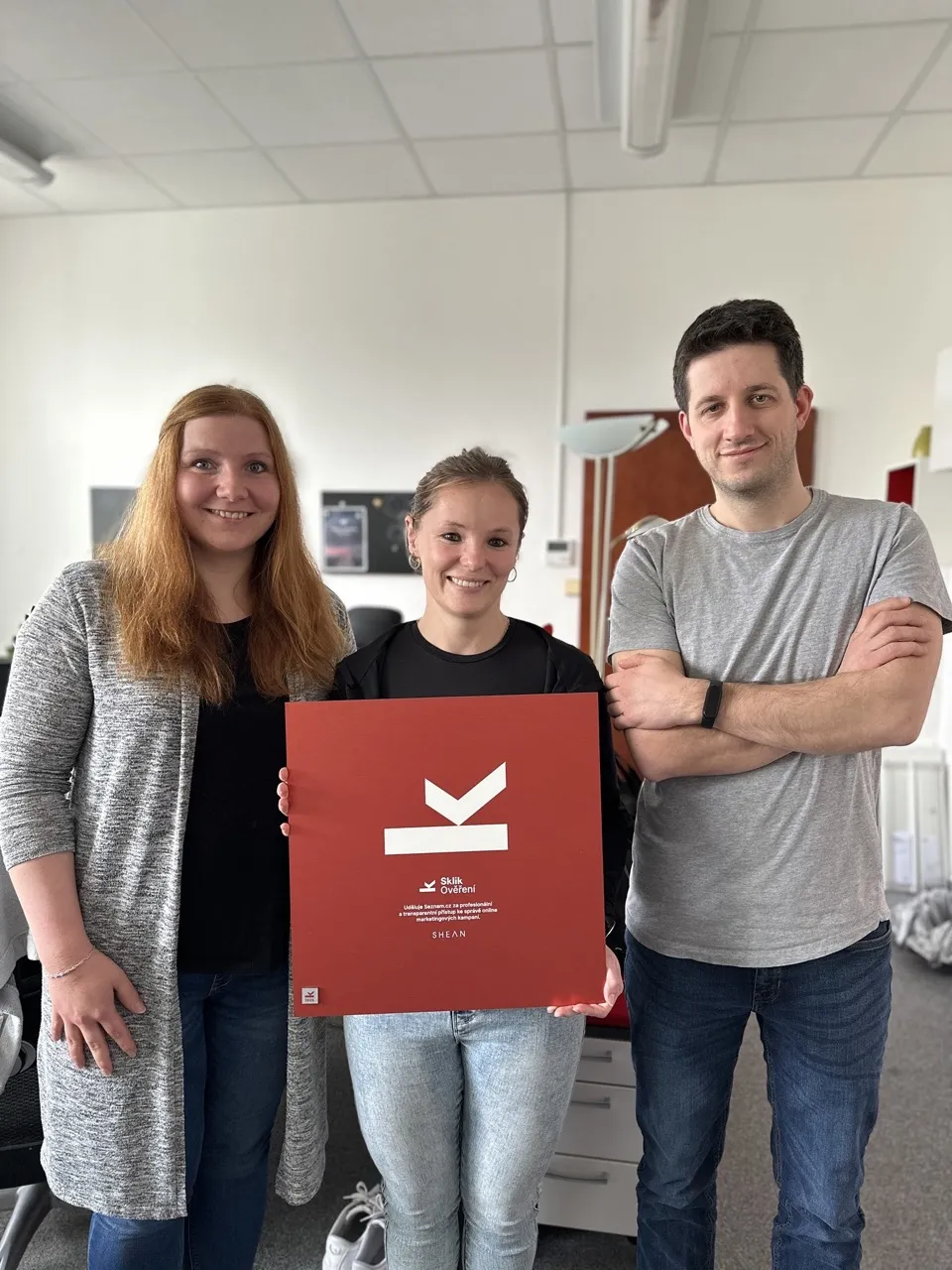 Tři usmívající se lidé v kanceláři drží červený plakát s bílým symbolem a nápisem Sklik Ověření.