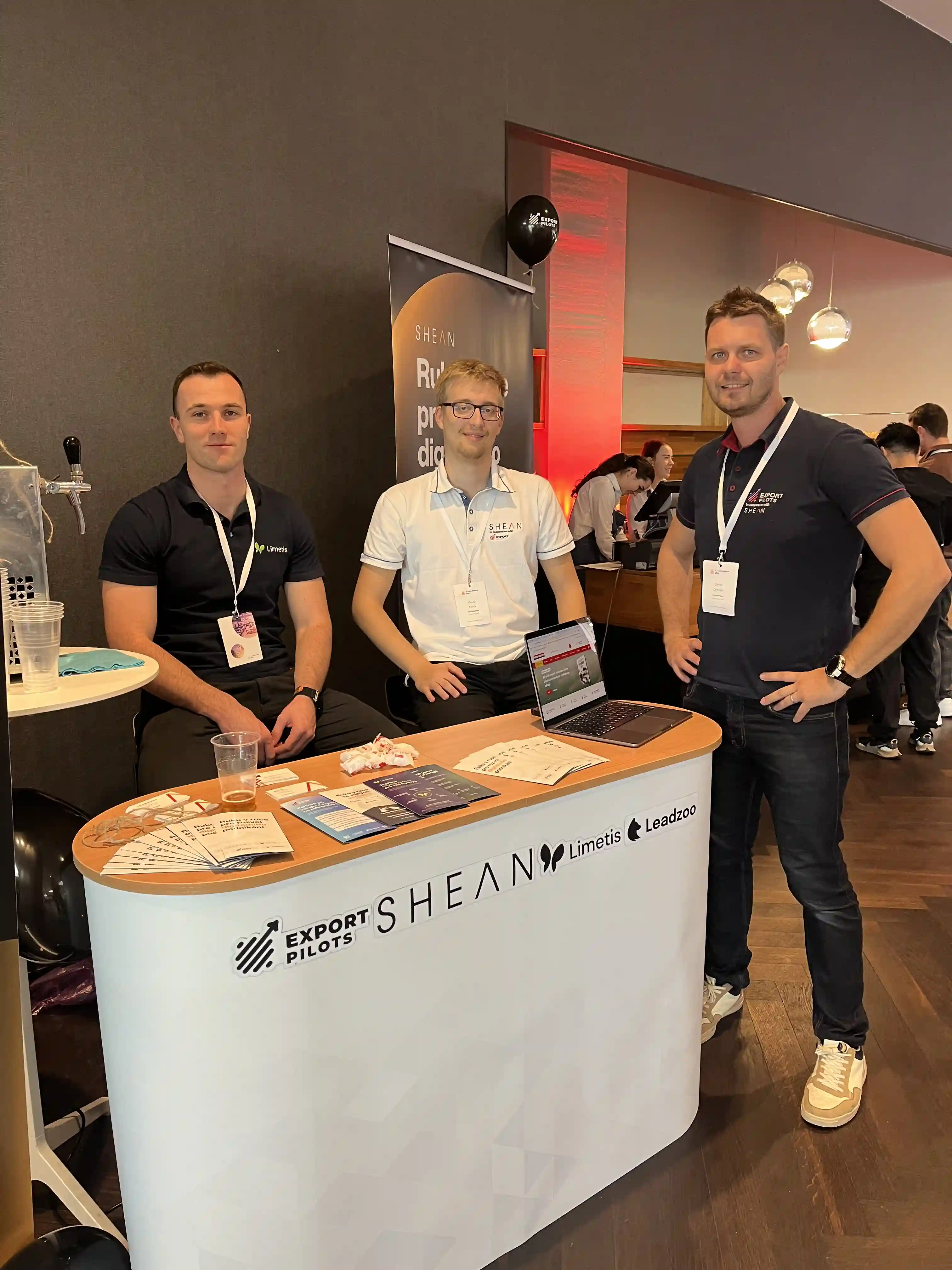 Tři muži se smějí a pózují u prezentačního stánku s logy Export Pilots, SHEAN, Limetis a Leadzoo, na stole letáky a otevřený laptop.