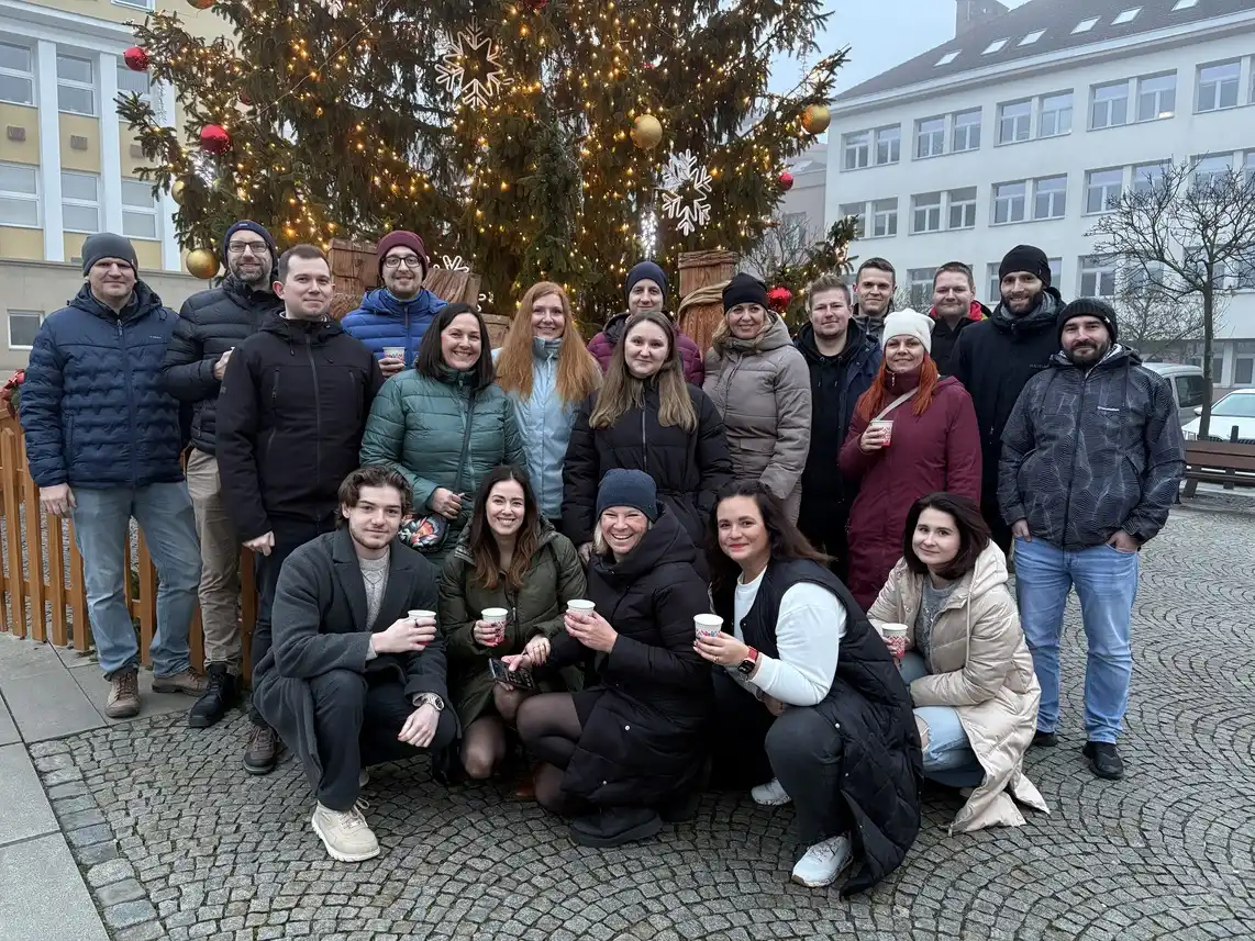 Skupina 21 lidí venku před osvětleným vánočním stromem, všichni v zimních bundách drží hrnky s nápoji.
