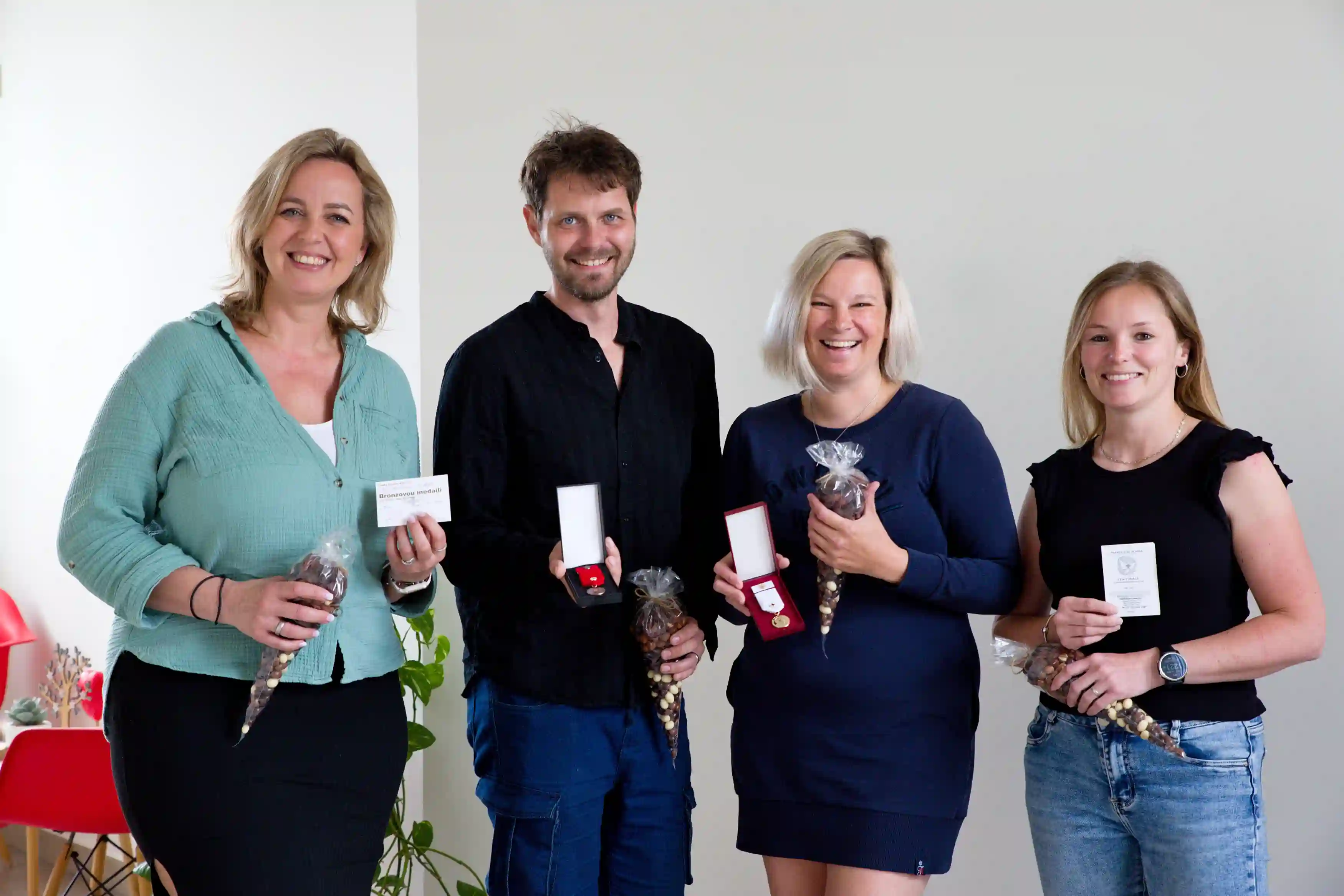 Čtyři usměvaví dospělí drží sáčky s oříšky a odznaky nebo ocenění ve svých rukou uvnitř místnosti.