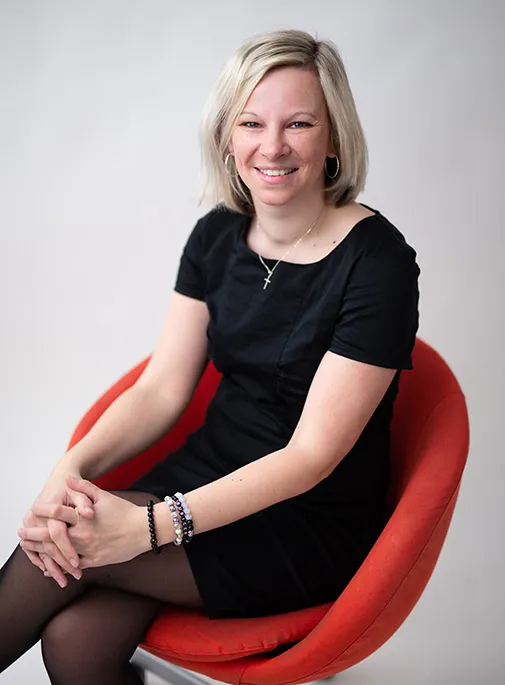 Usmívající se žena se světlými vlasy sedící v červeném křesle, oblečená v černých šatech a s náramky na zápěstí.