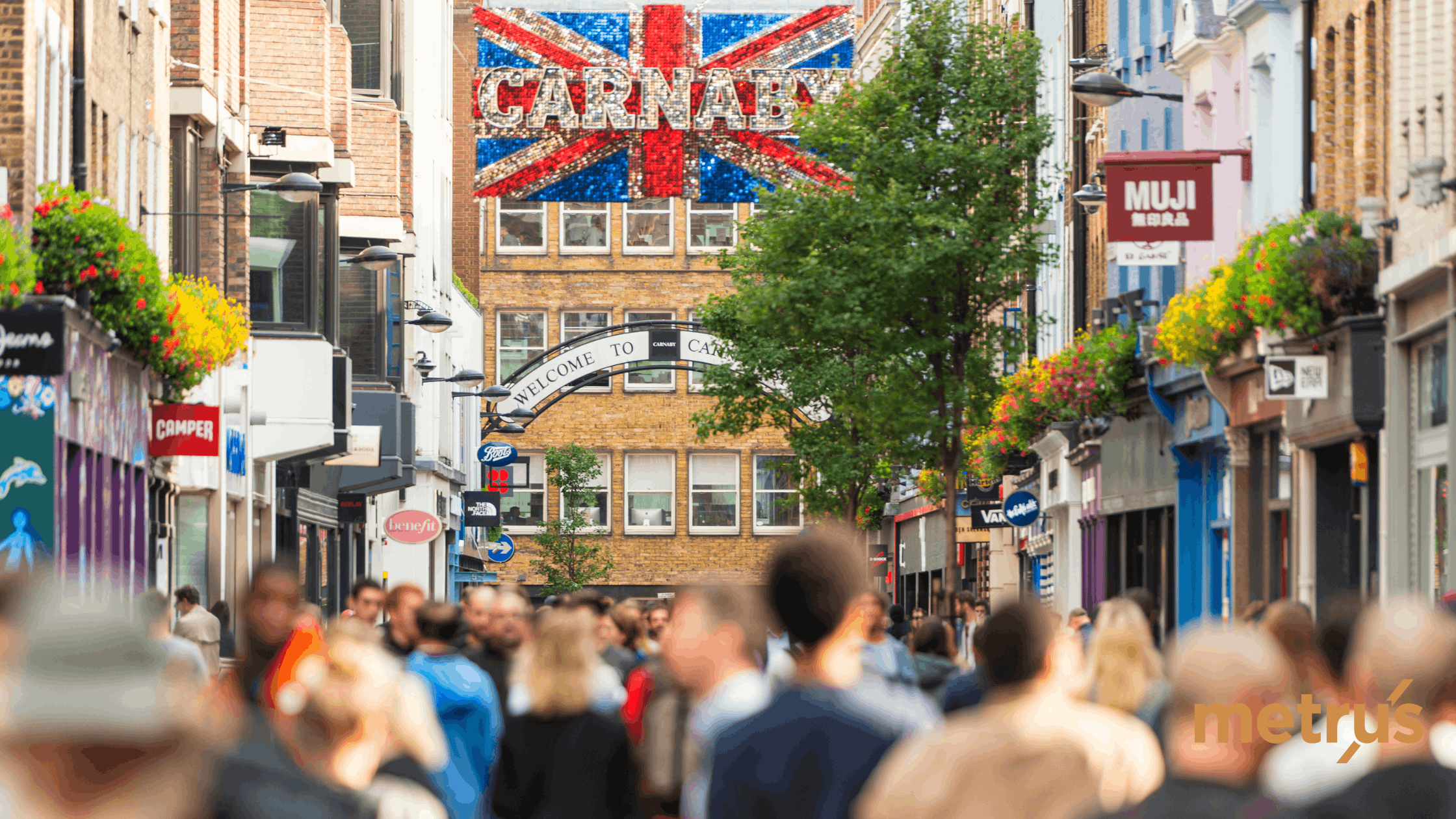 Crowded street in Carnaby, London with shops, hanging flower baskets, and a large Union Jack sign reading 'CARNABY'.