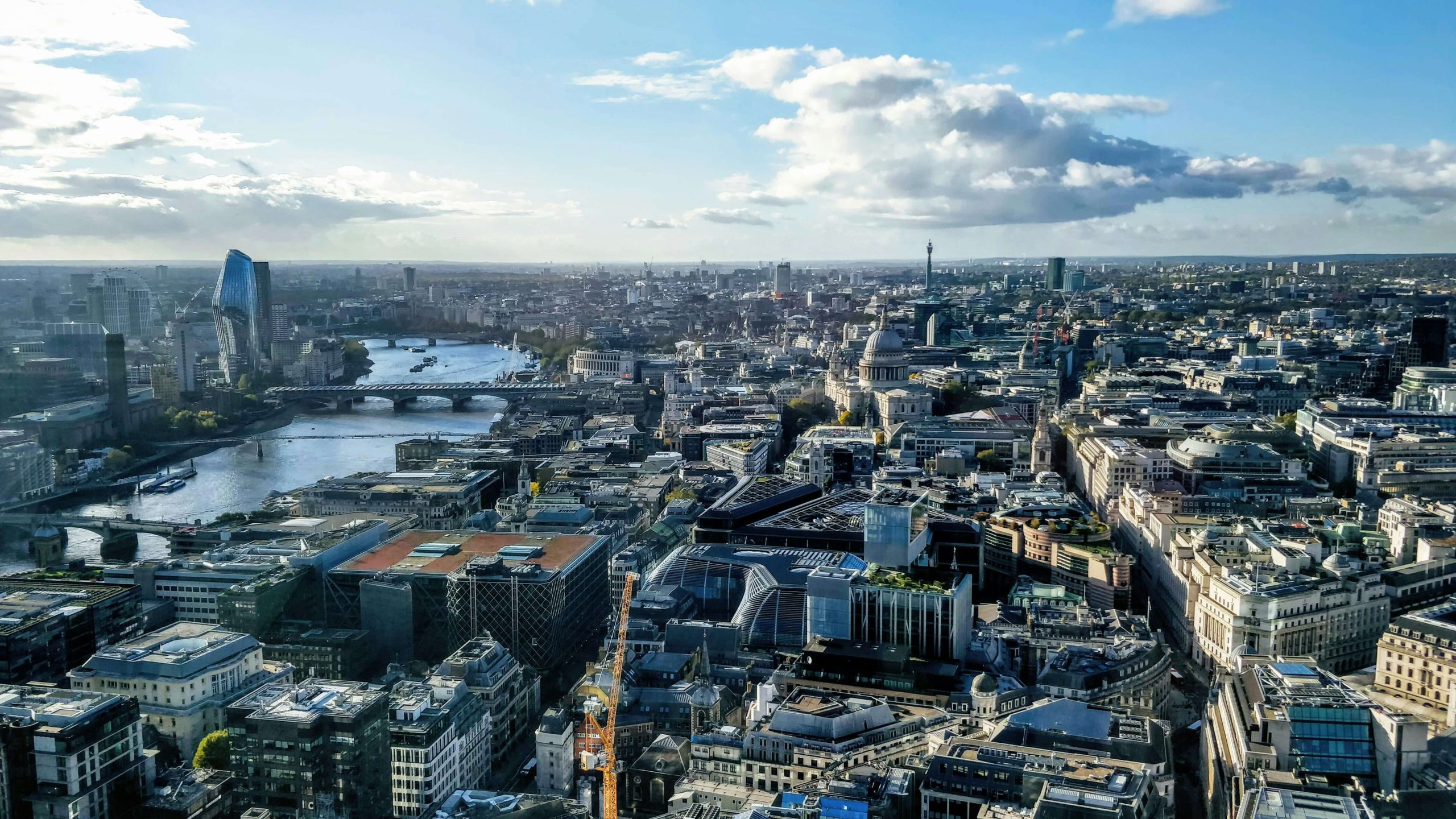 Aerial view of London skyline featuring the River Thames, multiple bridges, and St. Paul's Cathedral under a partly cloudy sky.