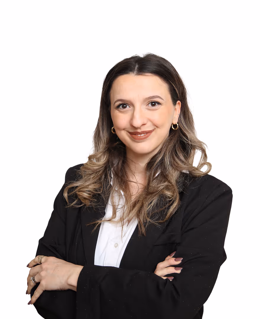 Lächelnde Frau mit lockigem Haar, schwarzem Blazer und weißem Hemd, die die Arme verschränkt hat.