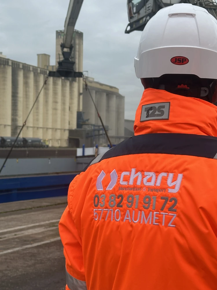 Employé portant un gilet fluorescent, un casque et des EPI sur un chantier de chargement d'une péniche de tubes de canalisation par une pelle de manutention de 60 tonnes