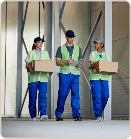 Three warehouse workers in safety vests and caps walking together, two carrying boxes and one holding a clipboard.