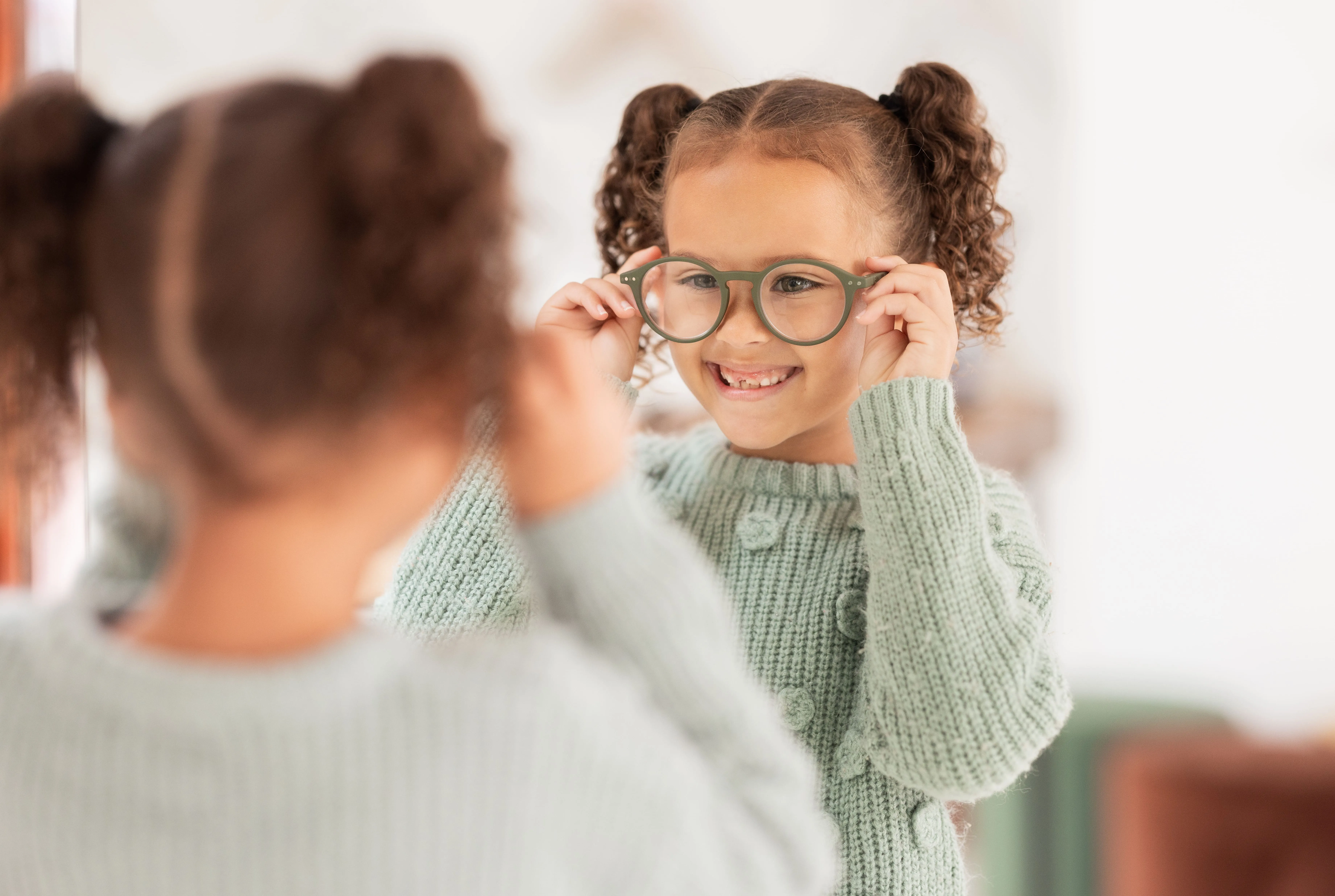 Young girl with curly hair and glasses smiling in mirror reflection