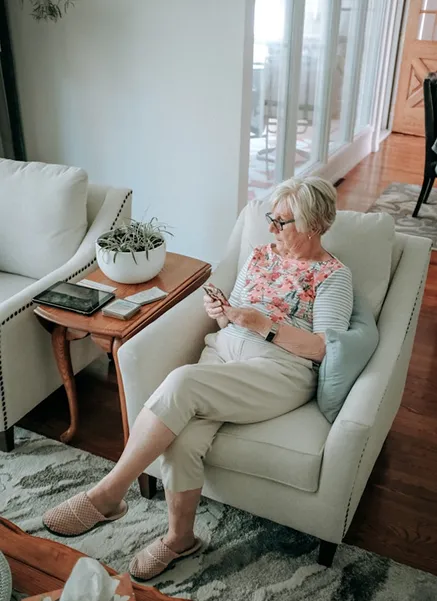 Elderly woman in glasses sitting on a cushioned armchair, looking at her smartphone in a cozy living room.