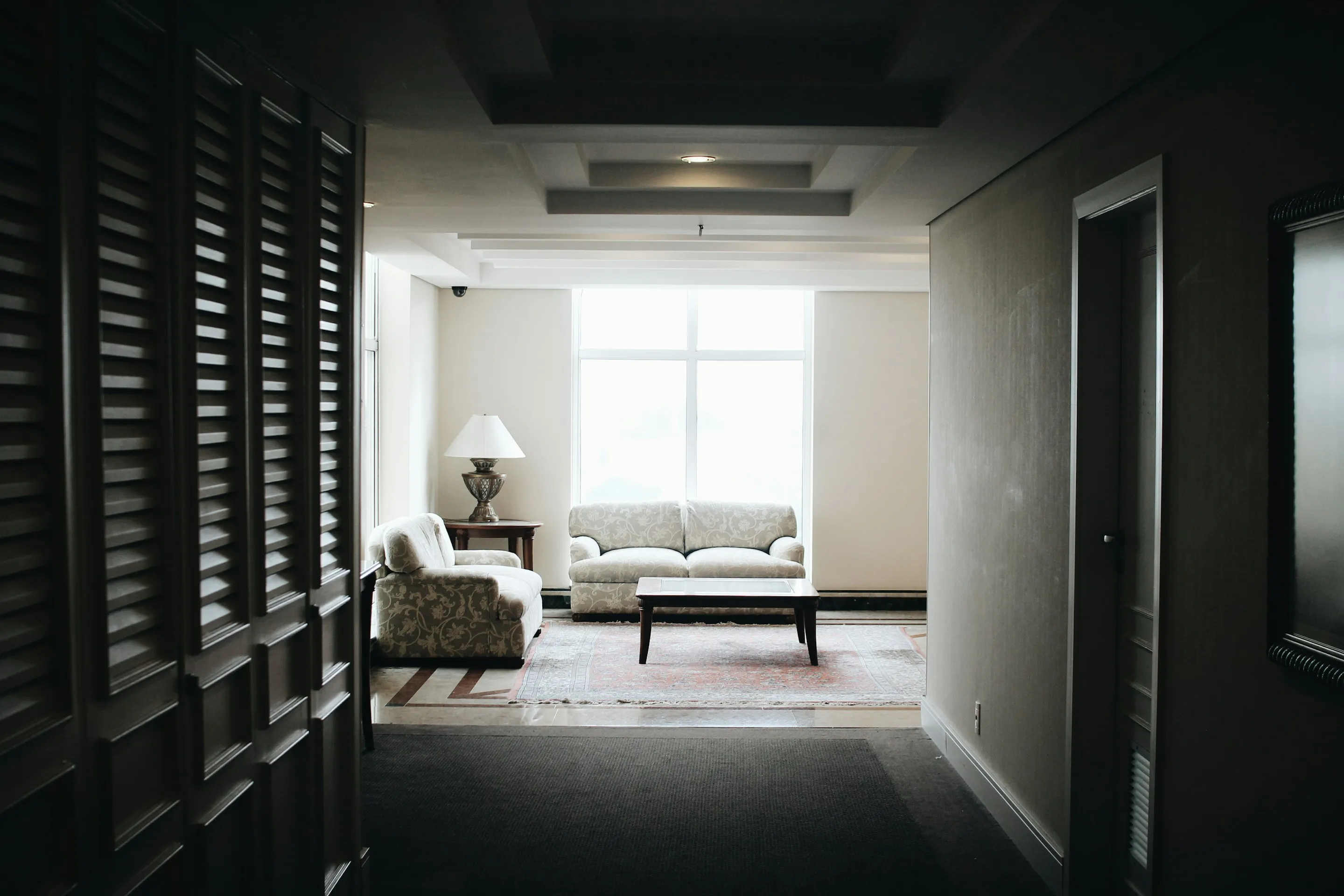Bright living area with floral-patterned sofas, a wooden coffee table on a rug, and a table lamp near a large window.