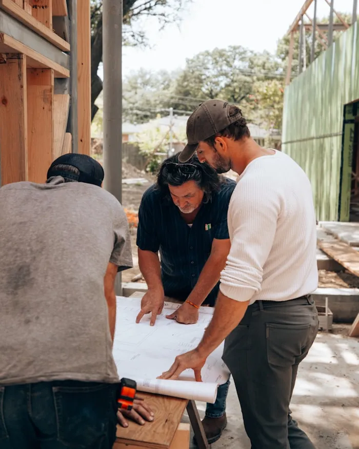 Three men reviewing architectural blueprints on a wooden table at a construction site.