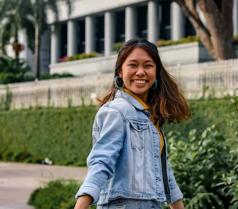 Smiling young woman wearing a light blue denim jacket and large hoop earrings outdoors near greenery and a building with columns.