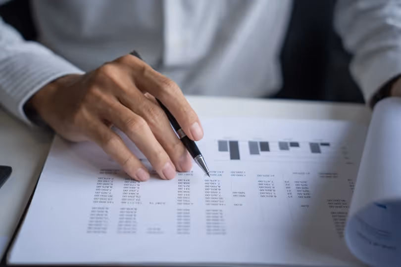 Person holding a pen and reviewing a printed financial document with charts and tables.