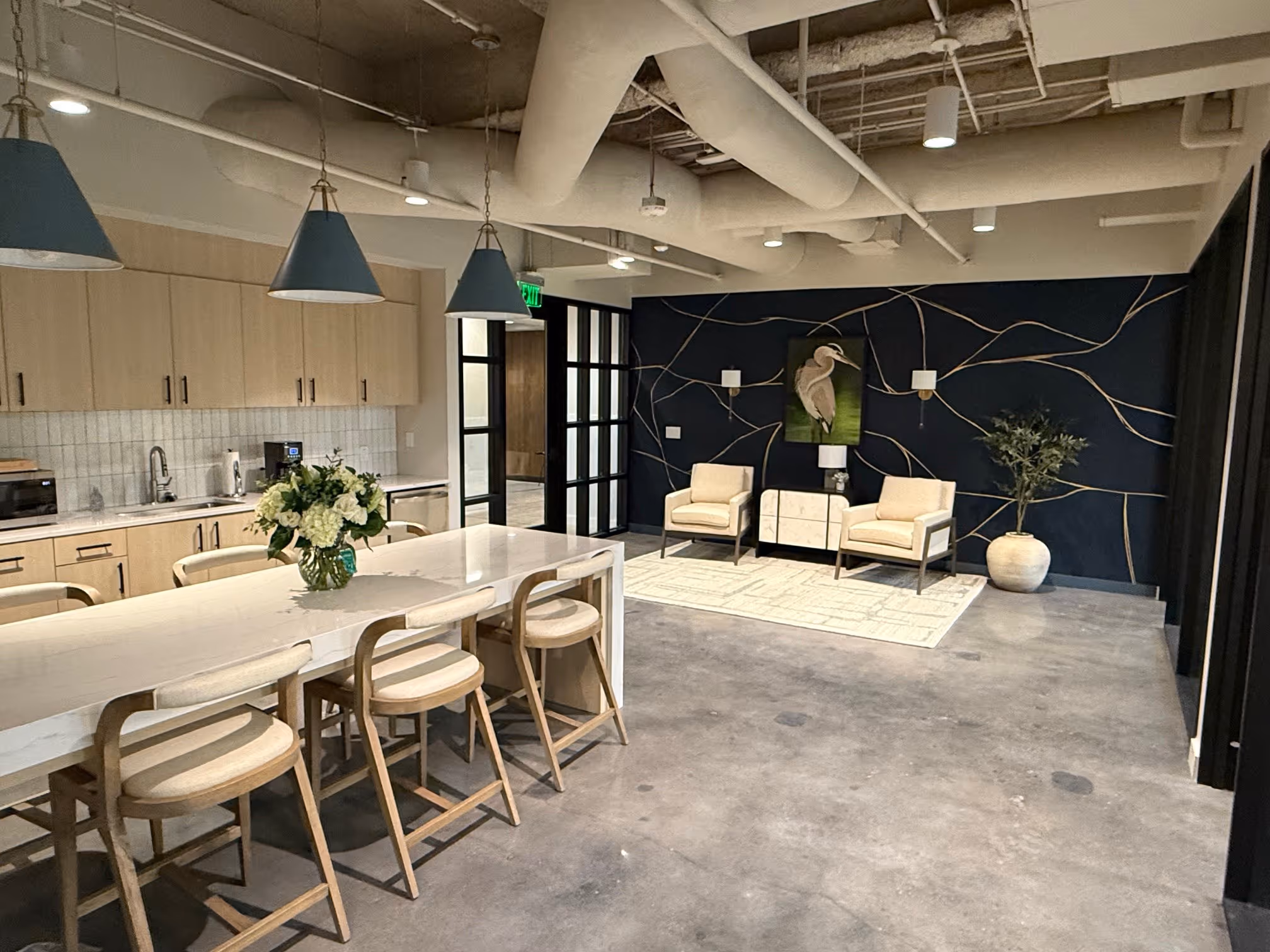 Modern office kitchen and lounge area with light wood cabinets, a white island with beige chairs, pendant lights, and a sitting area with cream armchairs, artwork of a bird, and a large potted plant.