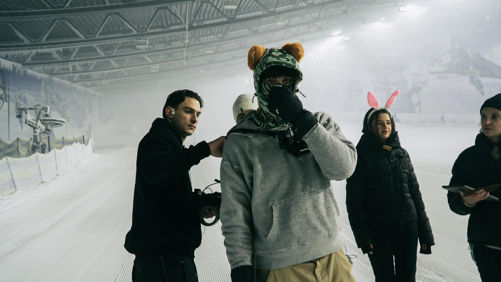 Fünf Personen in Winterkleidung stehen in einer schneebedeckten Halle, eine trägt eine Mütze mit Teddybär-Ohren, eine andere hat rosa Hasenohren.