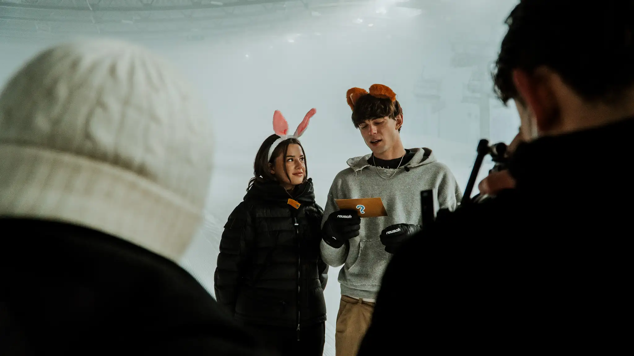 Junge Frau mit Hasenohren und junger Mann mit Bär-Ohren lesen gemeinsam eine Karte, während zwei Personen im Vordergrund beobachtend filmen.
