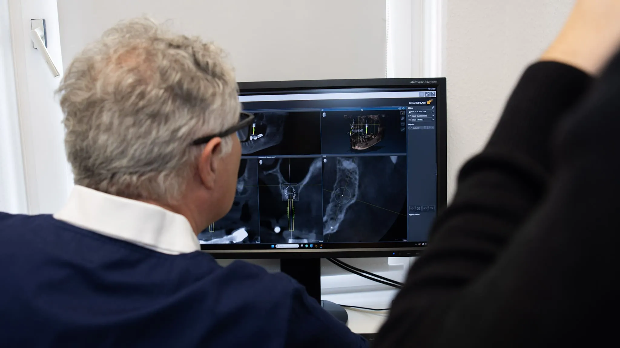 Zahnarzt betrachtet digitale Röntgenbilder und 3D-Implantatplanung auf einem Computerbildschirm.