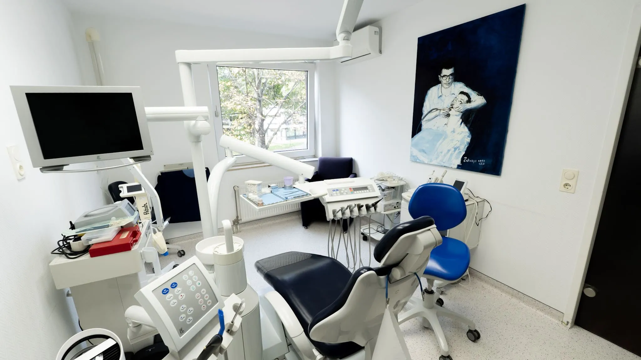 Modernes Behandlungszimmer einer Zahnarztpraxis mit Kunst an der Wand und Behandlungsstuhl vor großem Fenster.