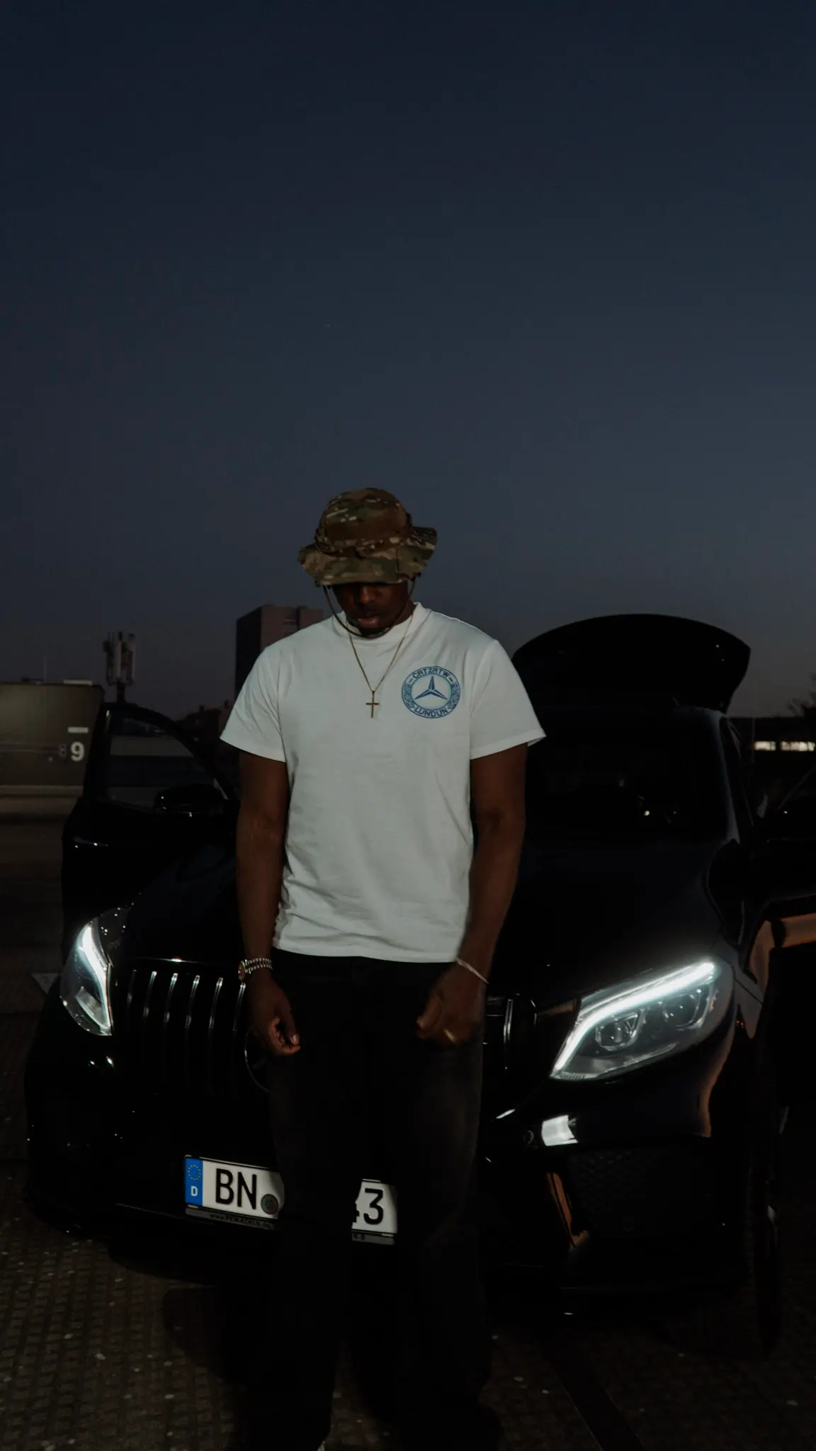 Nachtaufnahme auf einem Parkdeck – Mann mit weißem Shirt und Camo-Hut steht vor schwarzem Mercedes, aufgenommen von L’Étoile Visuals