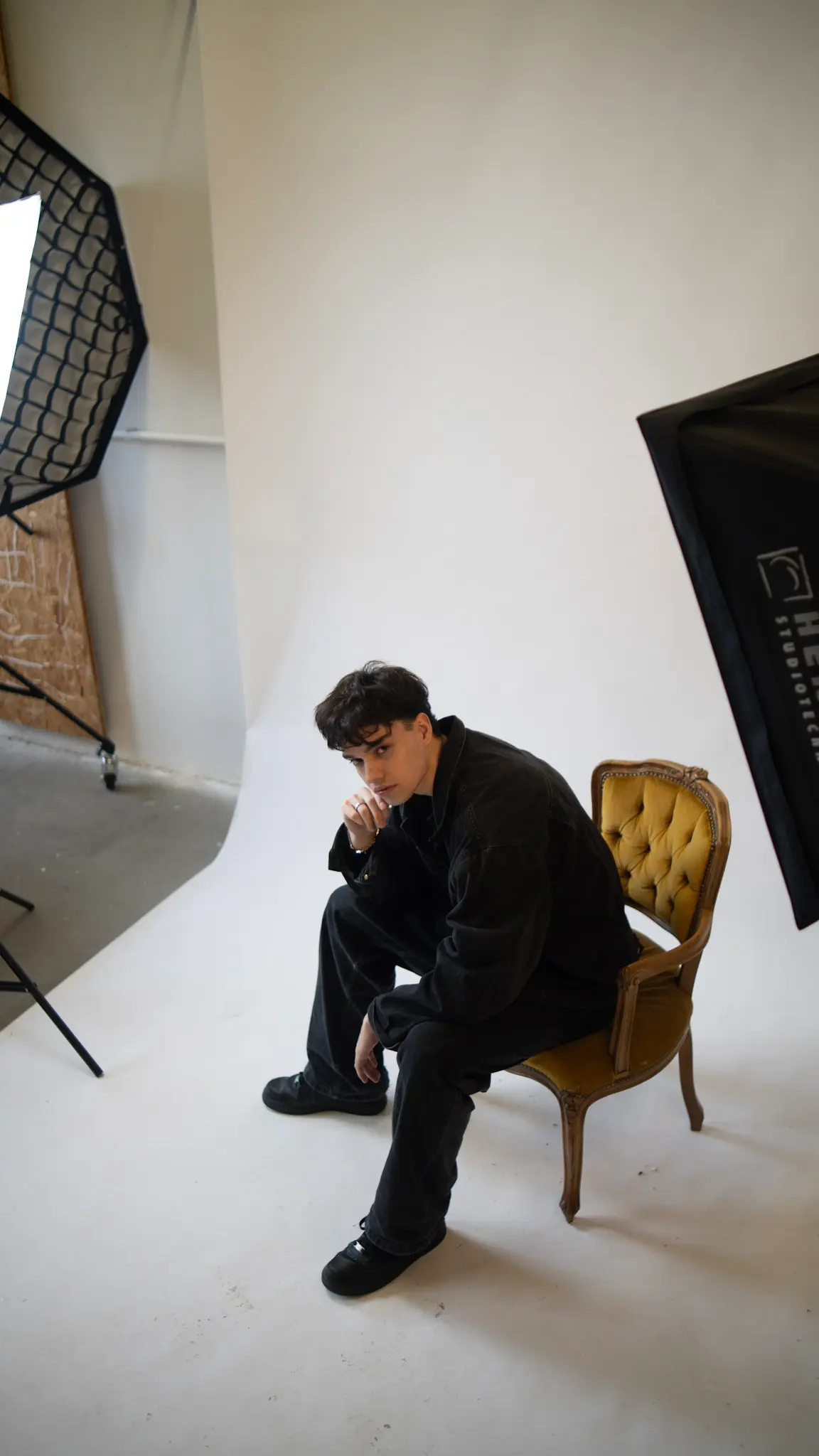 Studioaufnahme – junger Mann sitzt nachdenklich auf Vintage-Stuhl vor hellem Hintergrund, minimalistischer Fashion-Look, fotografiert von L’Étoile Visuals