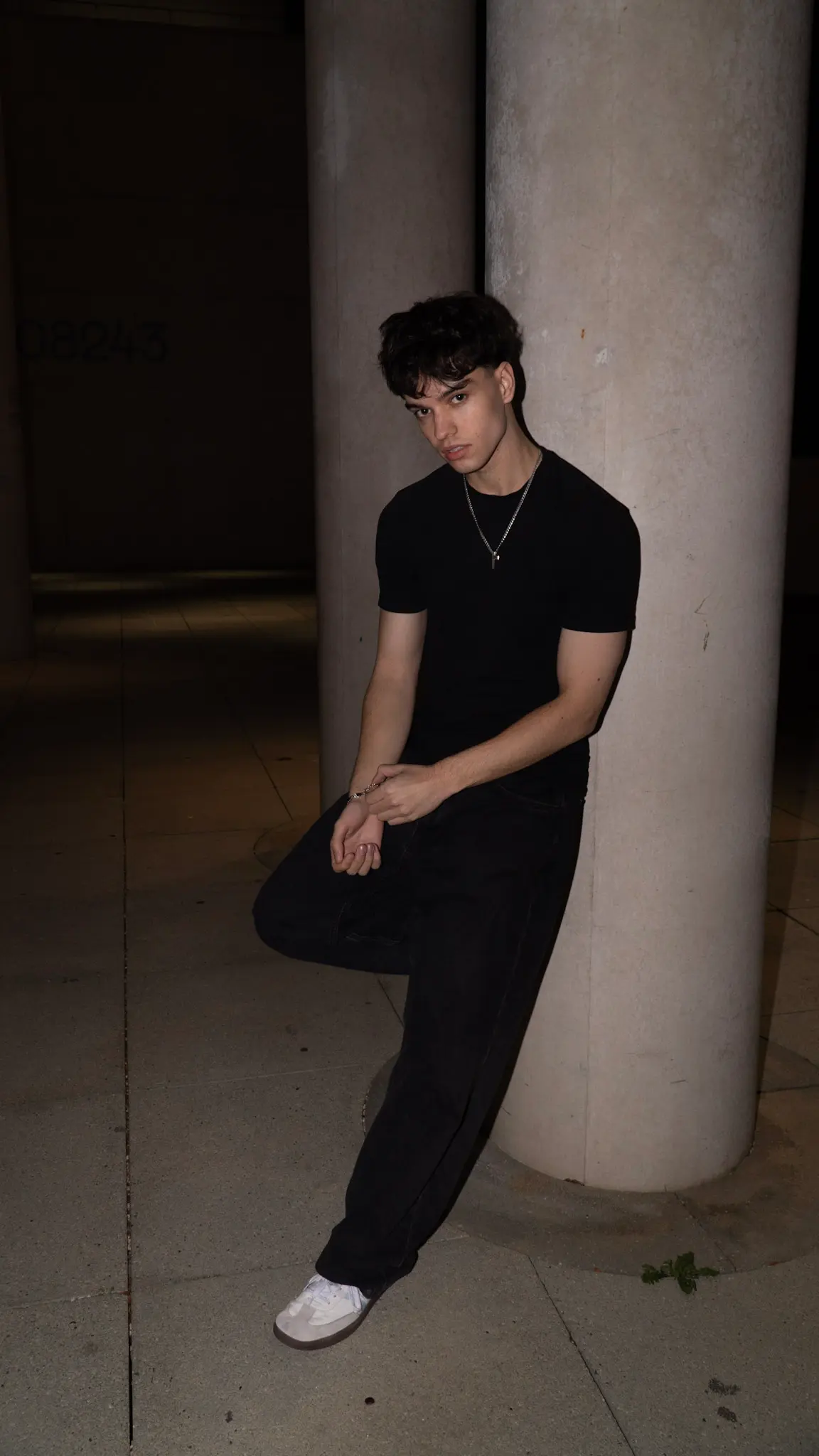 Nachtporträt eines jungen Mannes in schwarzem T-Shirt, lehnt an einer hellen Säule, minimalistischer Streetstyle-Look, fotografiert von L’Étoile Visuals