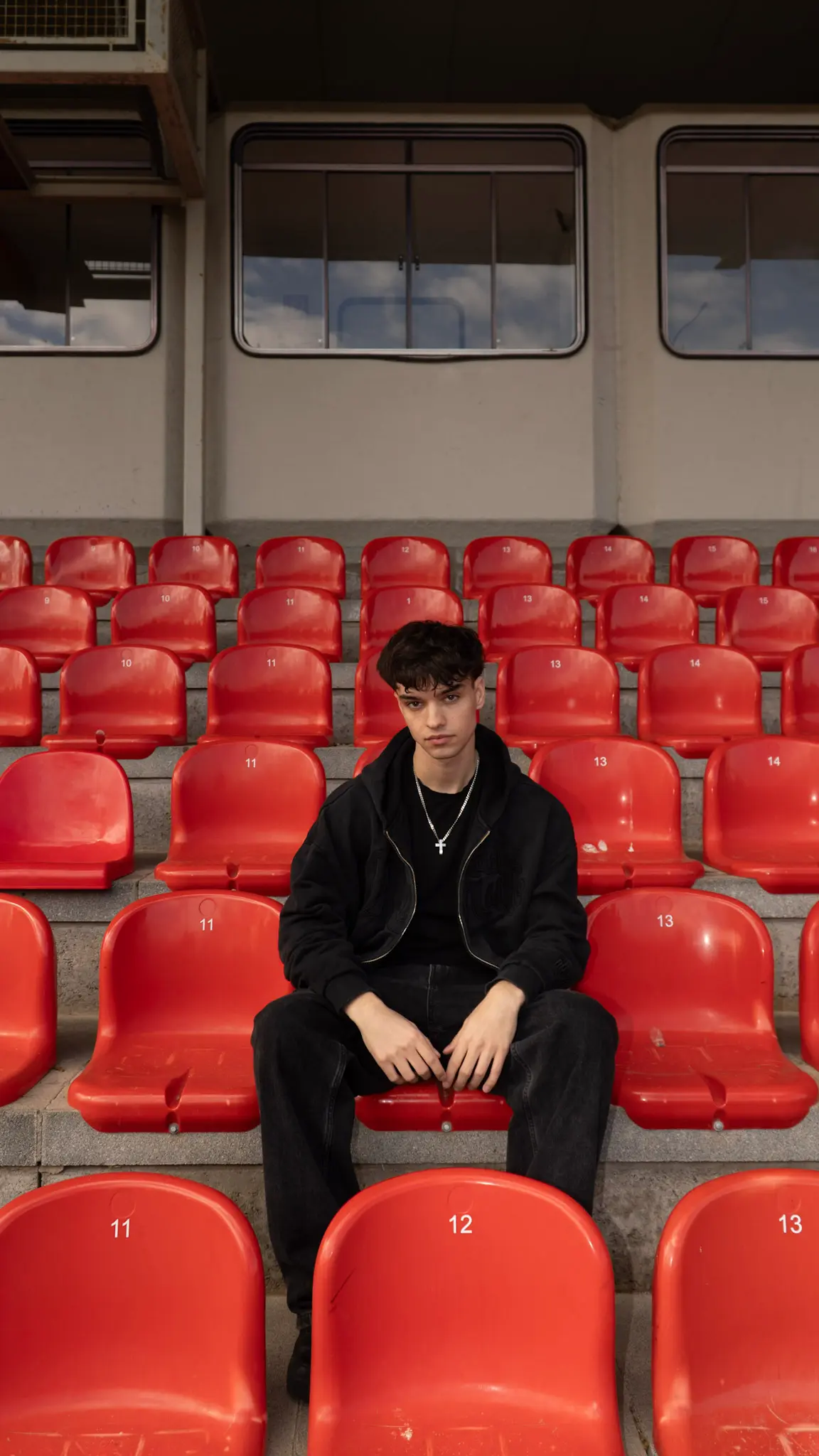 Editorial-Porträt eines jungen Mannes in schwarzem Outfit, sitzt in roten Stadionreihen, aufgenommen von L’Étoile Visuals
