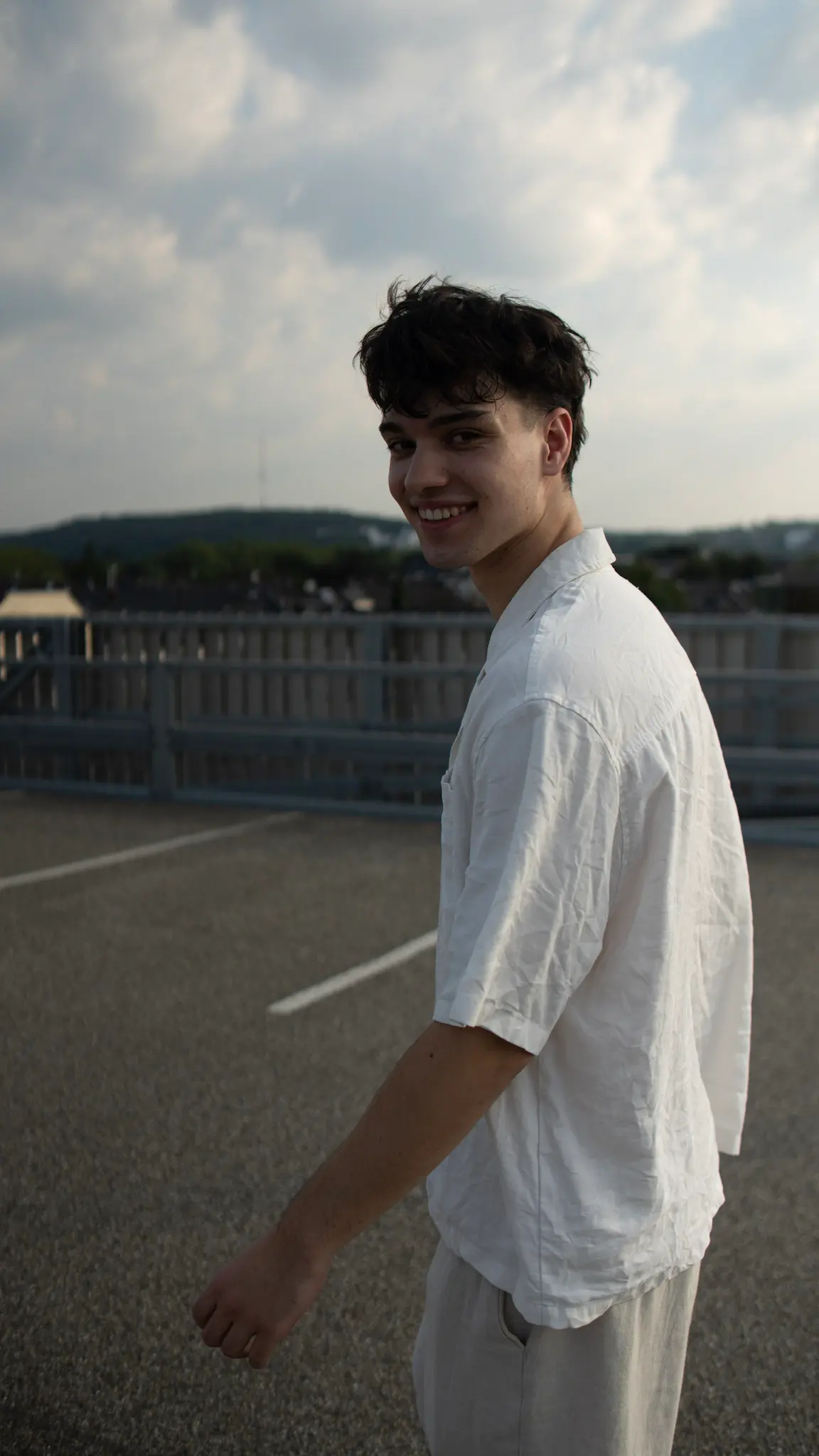 Natürliches Lifestyle-Porträt eines jungen Mannes im weißen Hemd auf einem Parkhausdach, lächelnd in die Kamera, fotografiert von L’Étoile Visuals
