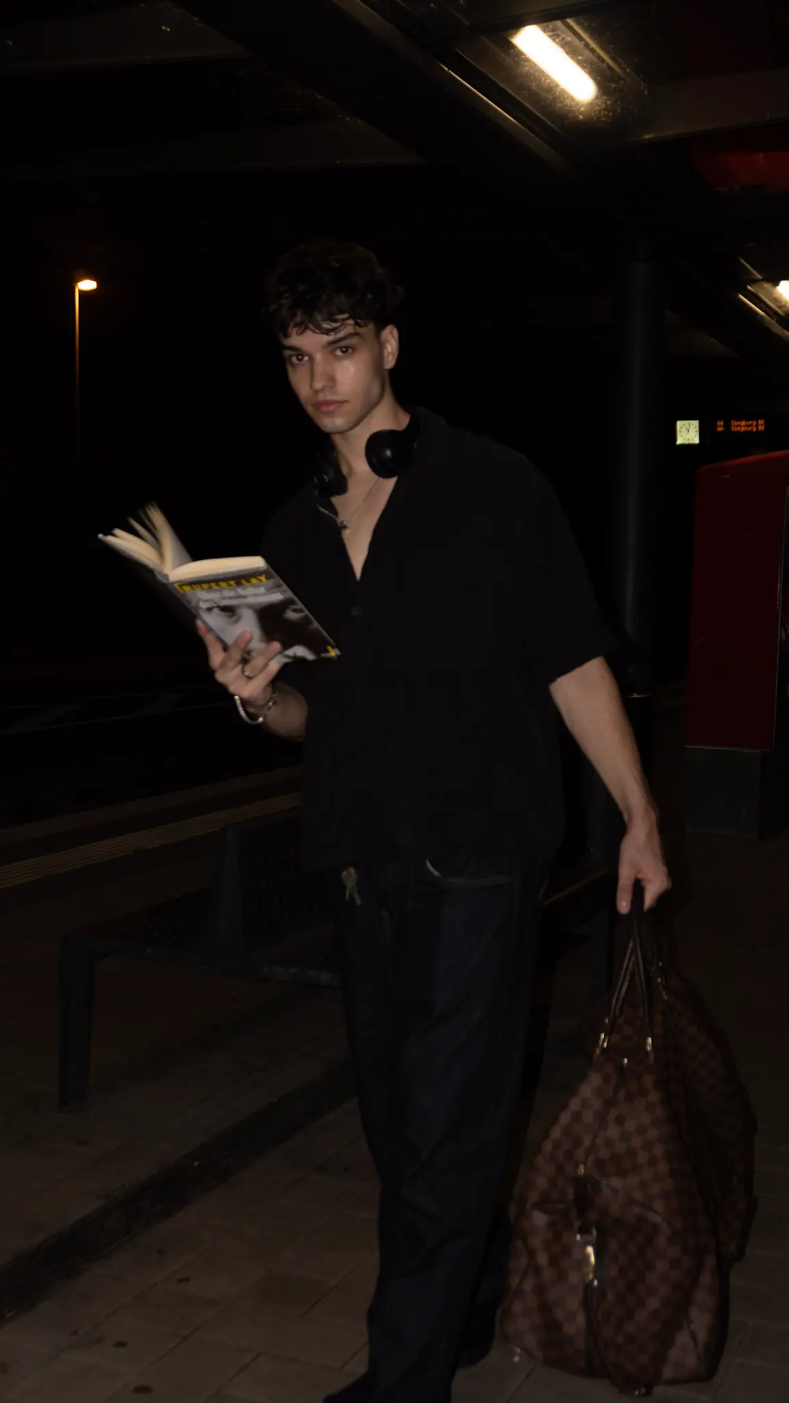 Urbanes Nachtporträt eines jungen Mannes, der ein Buch liest und eine Designer-Tasche trägt, in dunkler Kleidung am Bahnsteig, fotografiert von L’Étoile Visuals