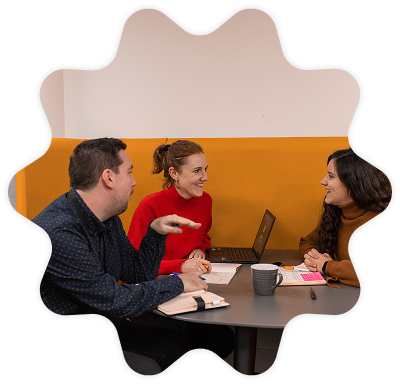 Three Penguin Books UK employees sitting in an orange booth and smiling whilst working on their laptops. 
