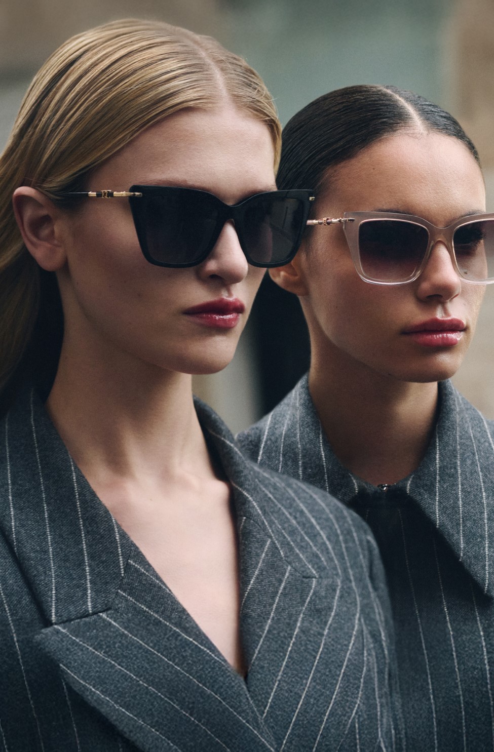 Two women wearing sunglasses and gray pinstriped blazers posing together outdoors.