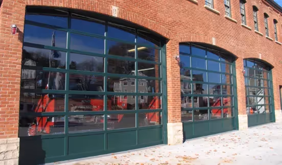 Brick building with large green-framed glass garage doors, likely a modern fire station or vehicle facility, with reflections of houses and trees in the glass.