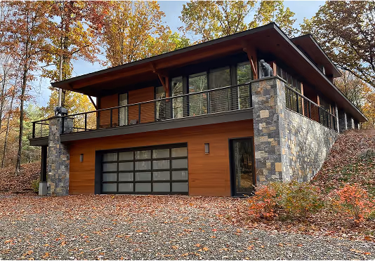 Modern two-story house surrounded by autumn foliage, featuring a glass-panel garage door, stone and wood exterior, and a balcony with black railing.