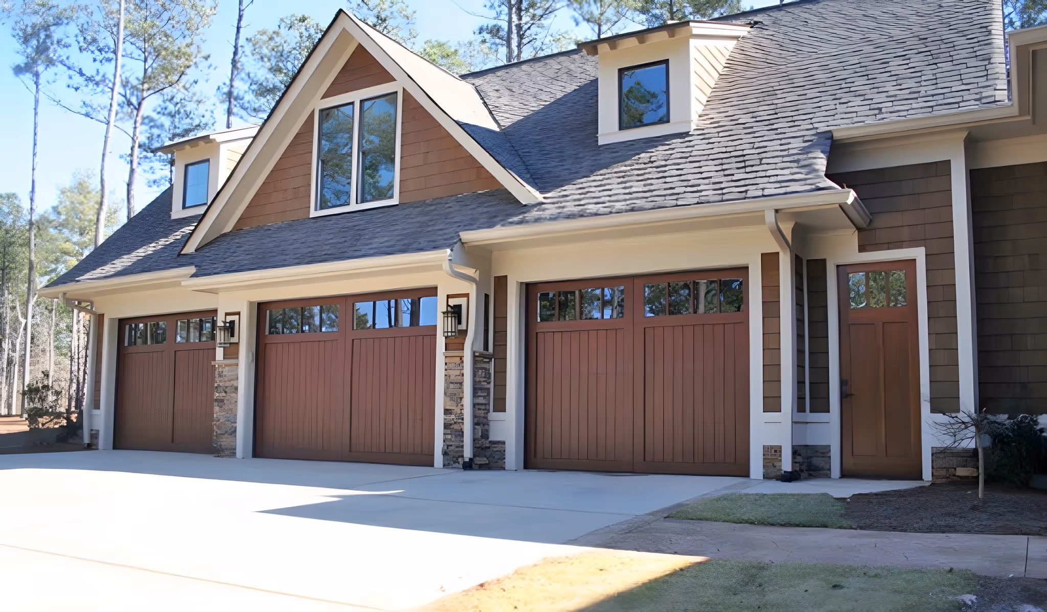 Traditional-style suburban home with three wooden garage doors featuring upper glass panels, stone accents, and a sloped shingle roof surrounded by tall trees.