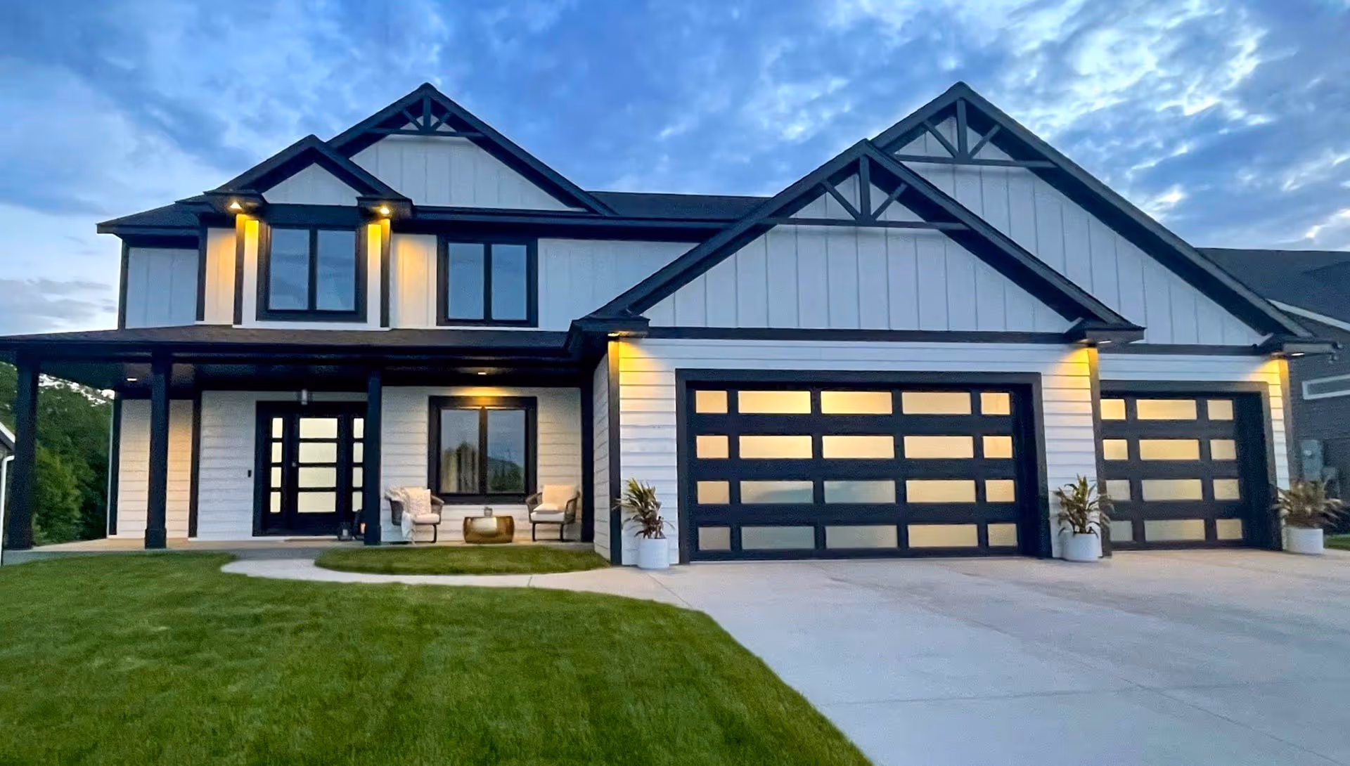 Modern two-story home with a white and black exterior, illuminated warm lighting, and two large glass-panel garage doors, situated on a well-manicured lawn at dusk.