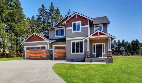 Two-story suburban home with gray siding, stone accents, and wooden garage doors, set against a backdrop of tall evergreen trees and a bright blue sky.
