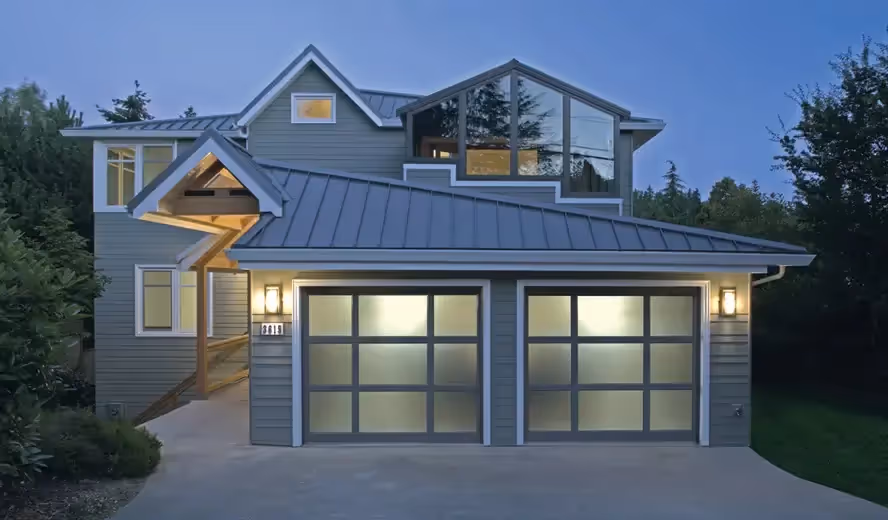 Modern two-story home with a sleek gray metal roof, large windows, and two frosted glass garage doors, softly illuminated in the evening light.