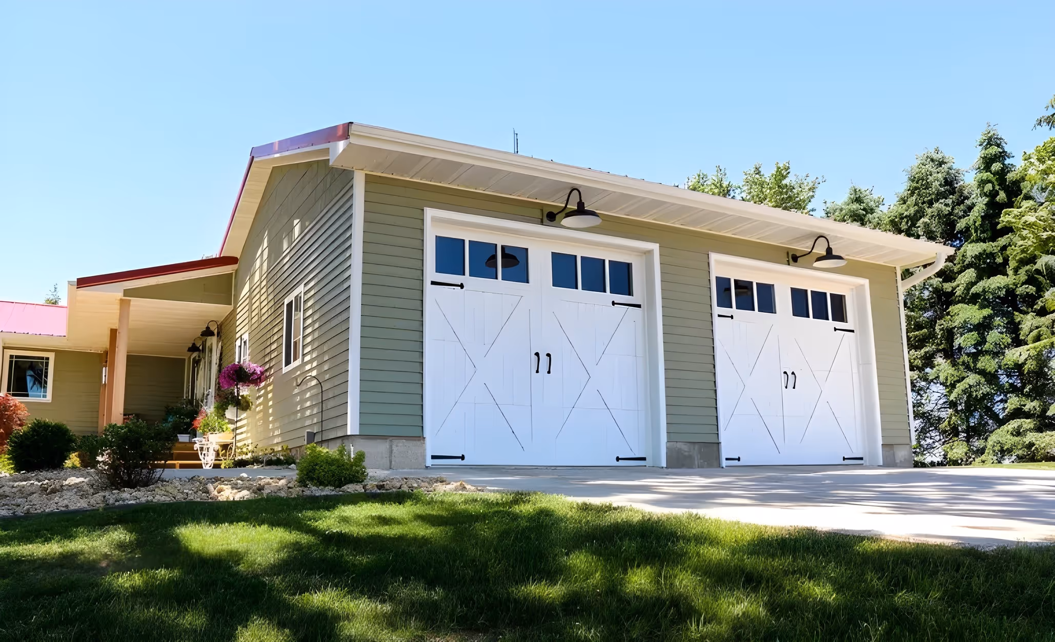 Modern farmhouse garage with two white barn-style doors, black hardware, and green siding. Red roof and simple landscaping.